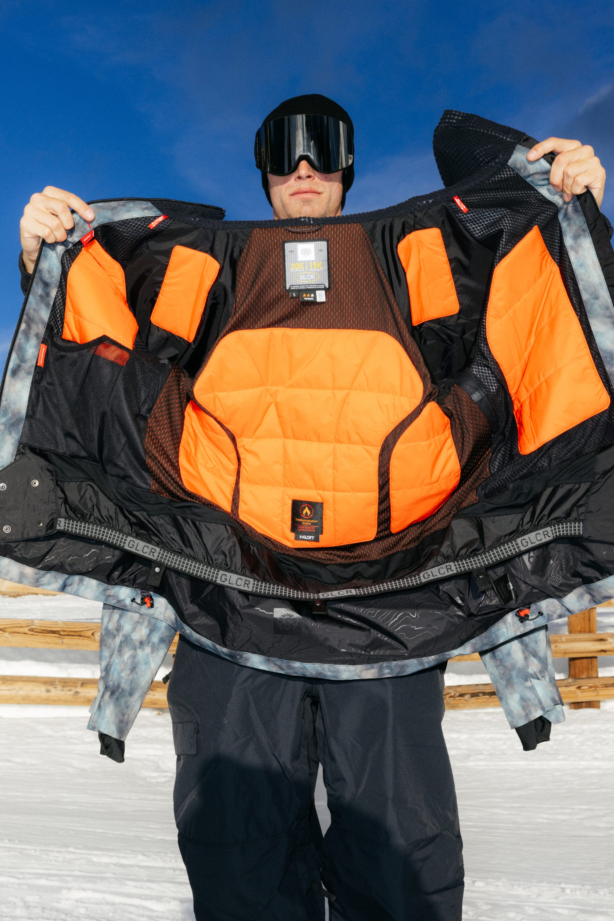 A person in snow gear holds open the 686 Men’s Hydra™ Thermagraph® Jacket by 686, revealing its bright orange, padded interior lining. The background features a snowy landscape and a wooden fence under a blue sky.