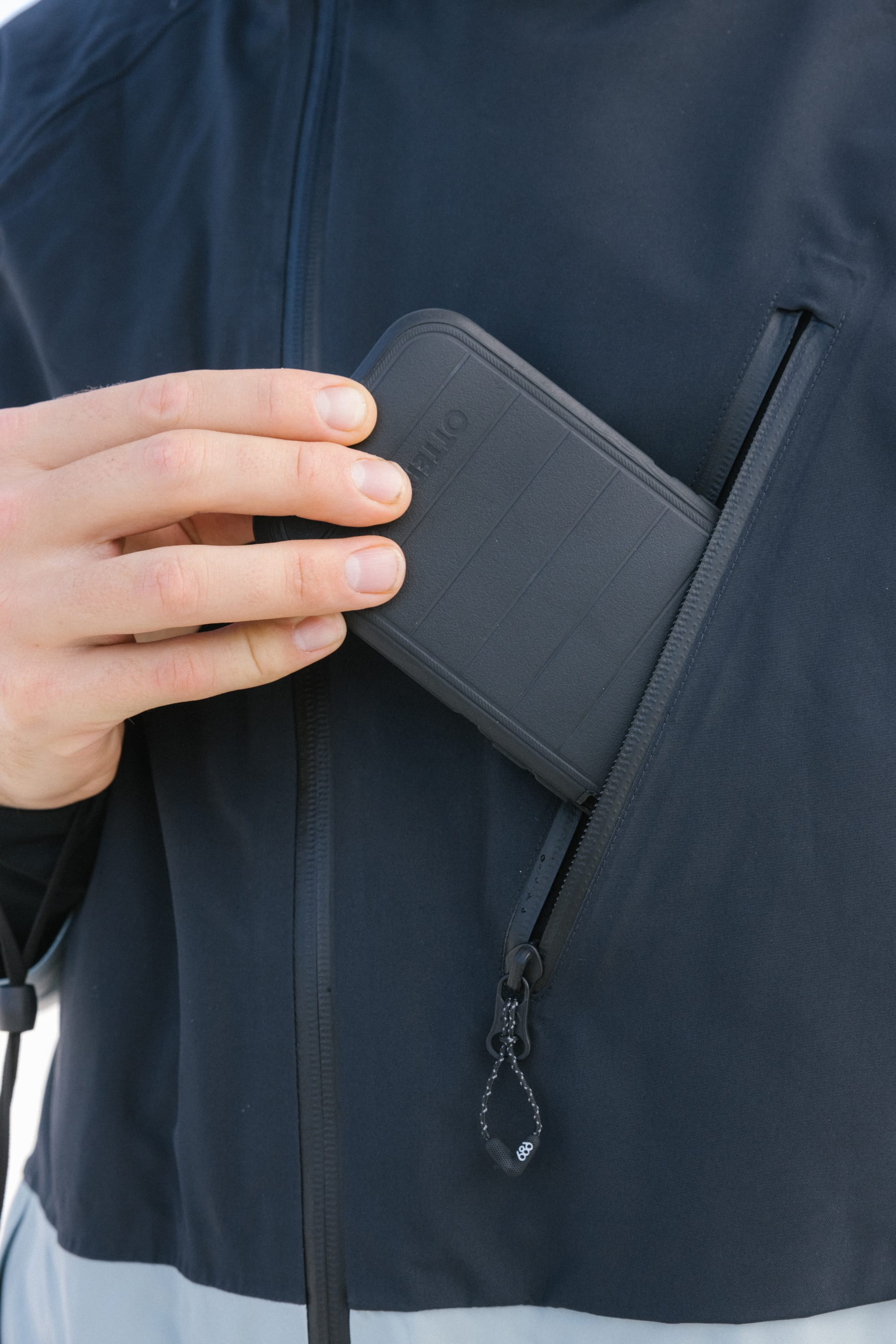 A person slips a black, rectangular device into the zippered chest pocket of the 686 Men's Hydra™ Thermagraph® Jacket, revealing only their hand, the pocket, and part of the jacket’s body-mapped lining.