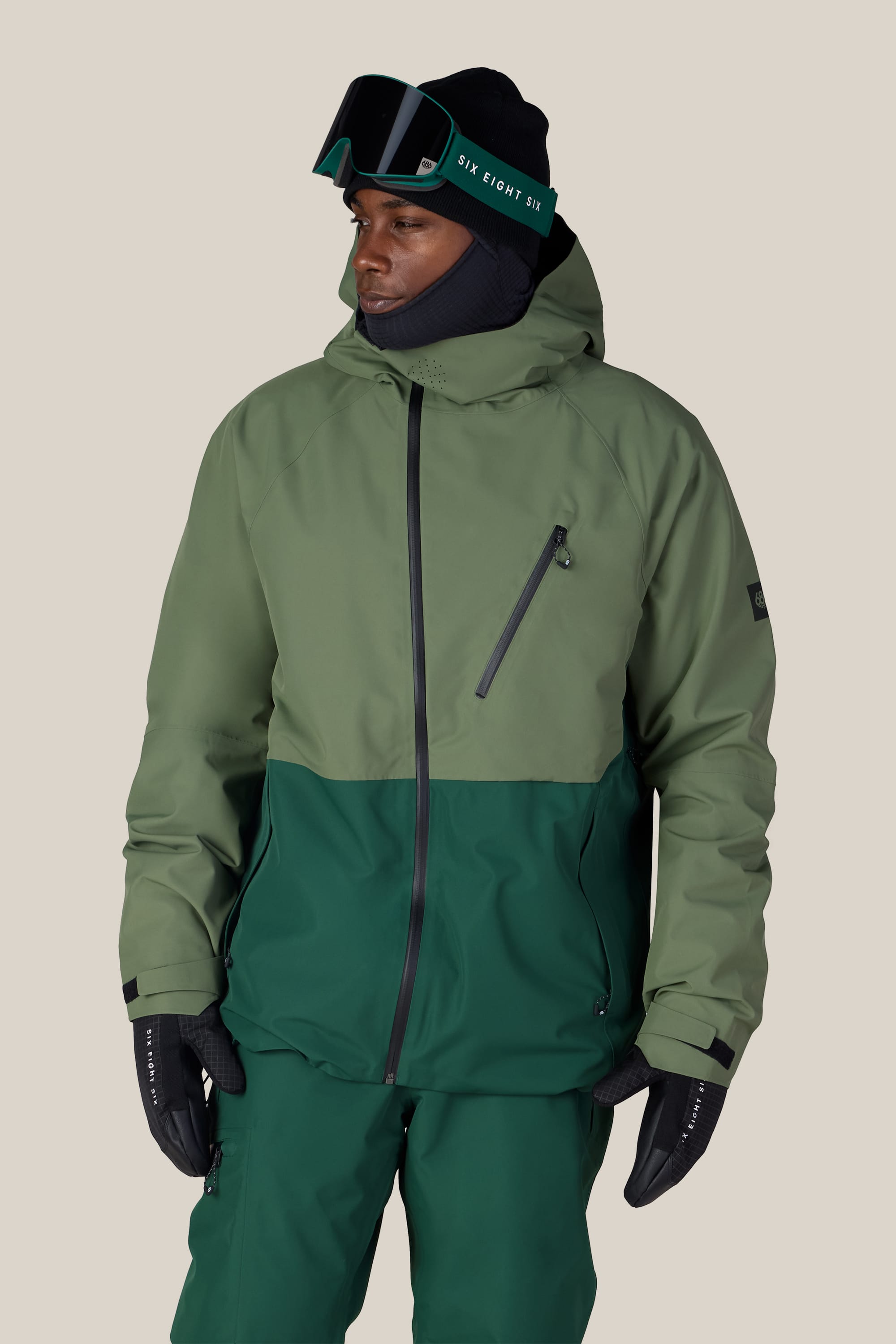 A person wearing the 686 Men’s Hydra™ Thermagraph® Jacket by 686, styled in shades of green with matching green pants, black gloves, a black balaclava, and ski goggles, stands against a plain beige background.