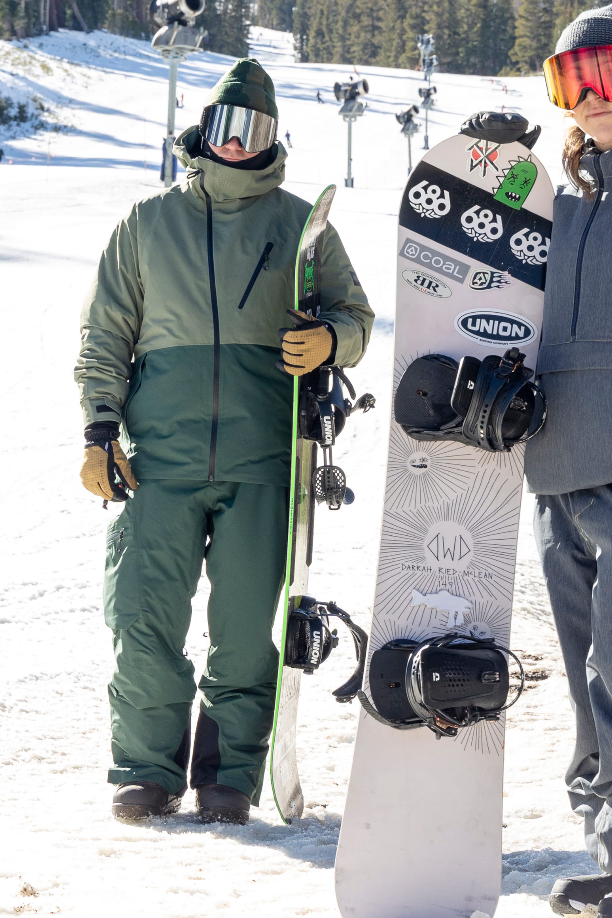 Two people dressed in 686 Men’s Hydra™ Thermagraph® Jackets stand on snowy ground holding snowboards. Ski lifts and trees are visible in the background on a sunny day.
