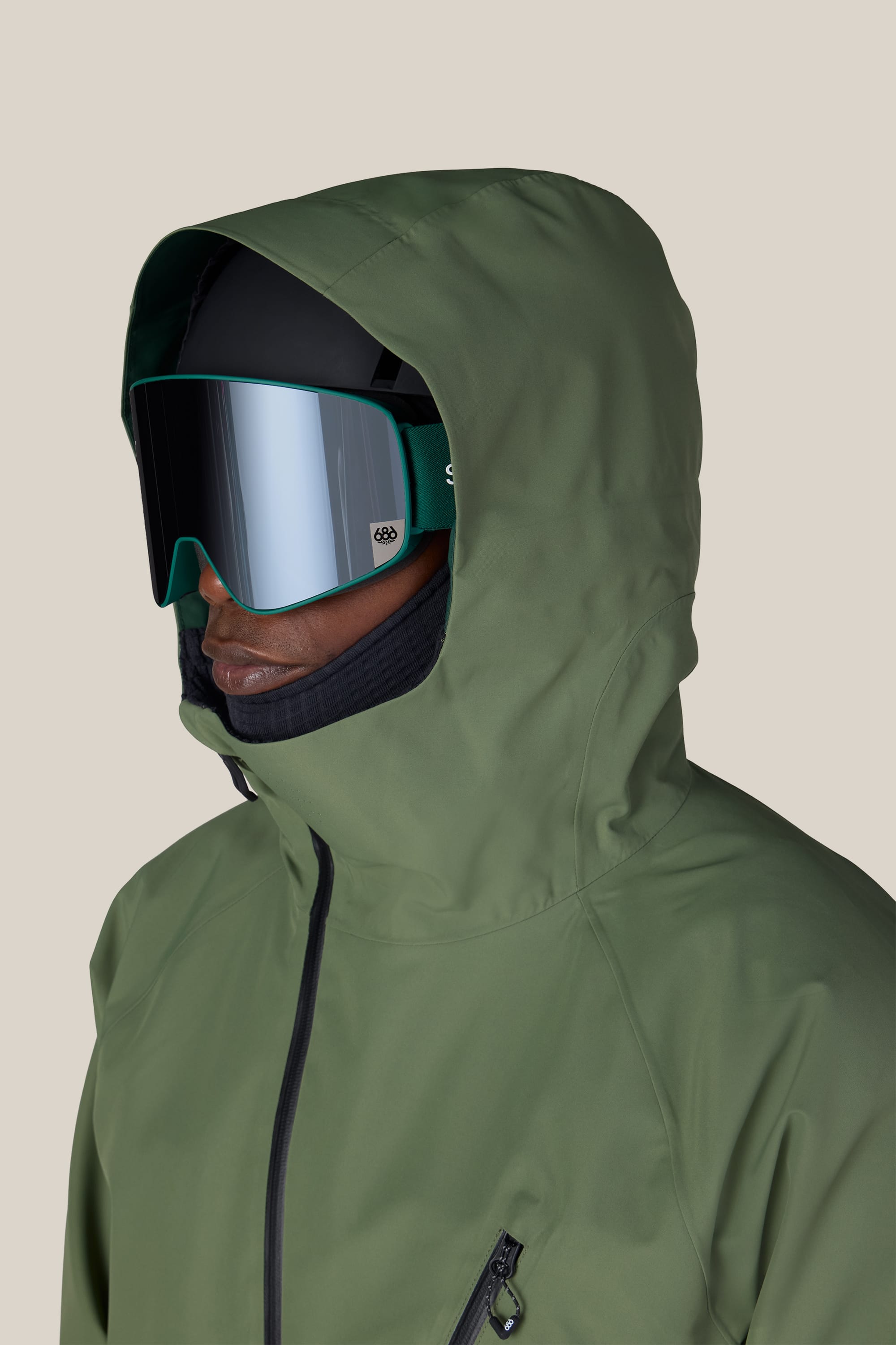 A person wearing the 686 Men’s Hydra™ Thermagraph® Jacket in green, along with black ski goggles and a black helmet, facing left against a plain light background.