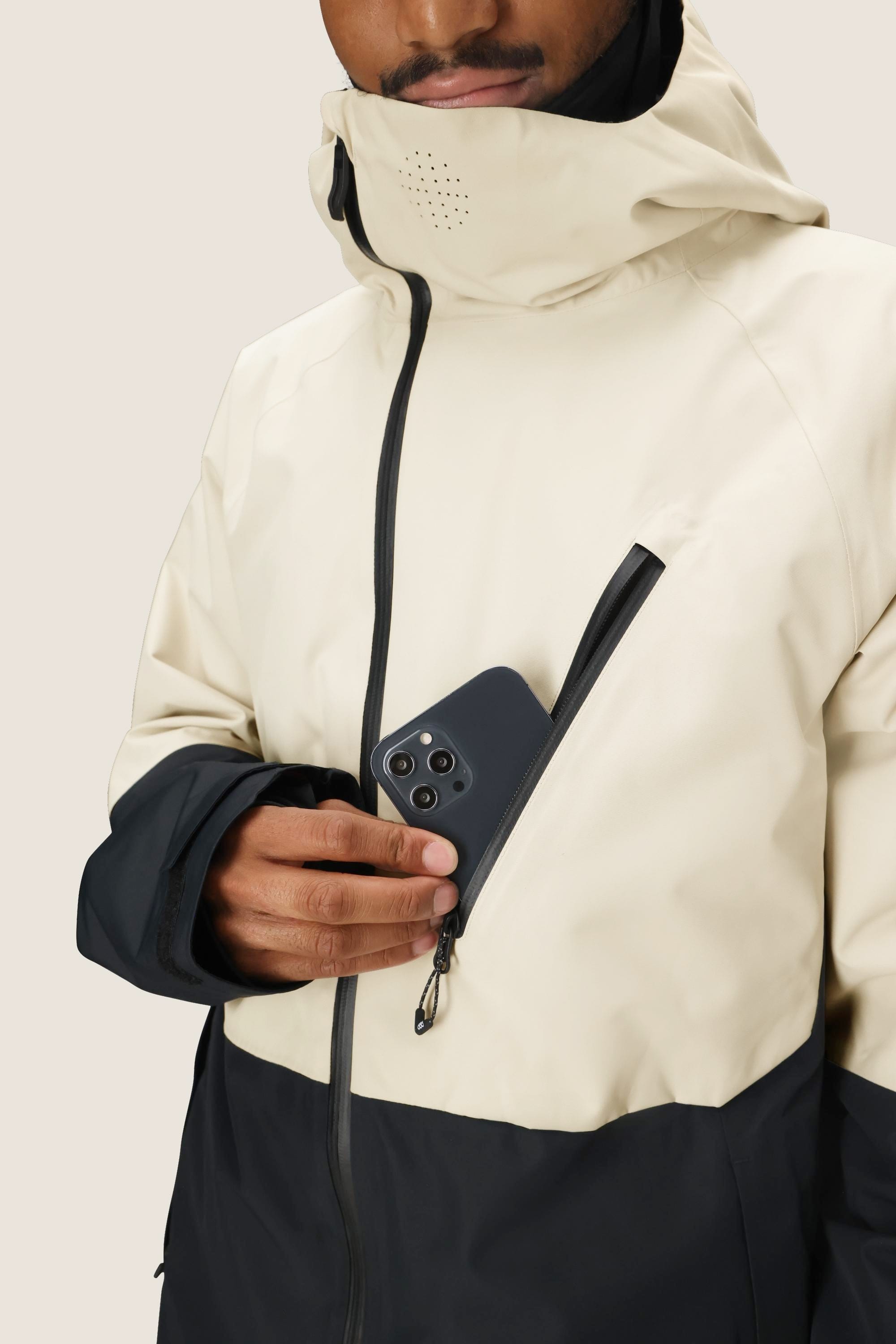 Wearing the 686 Men's Hydra™ Thermagraph® Jacket, featuring a hood and high collar, someone slips a smartphone into the chest pocket. The beige and black 686 jacket offers body-mapped lining for versatile comfort.