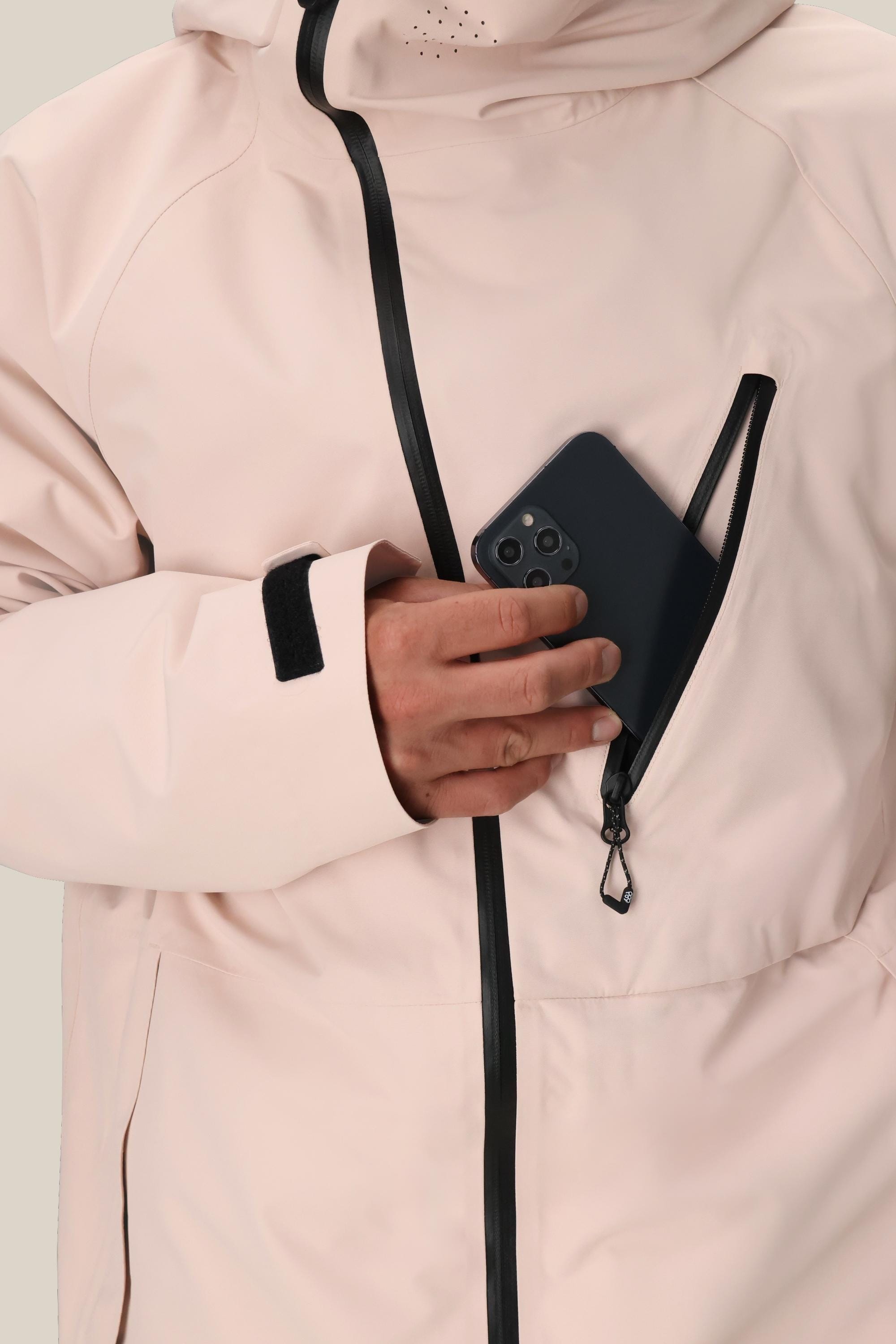 A person wearing the 686 Men's Hydra™ Thermagraph® Jacket in light pink zips a black smartphone into the chest pocket. This best seller from 686 features black zippers, body-mapped lining, and a Velcro sleeve strap.