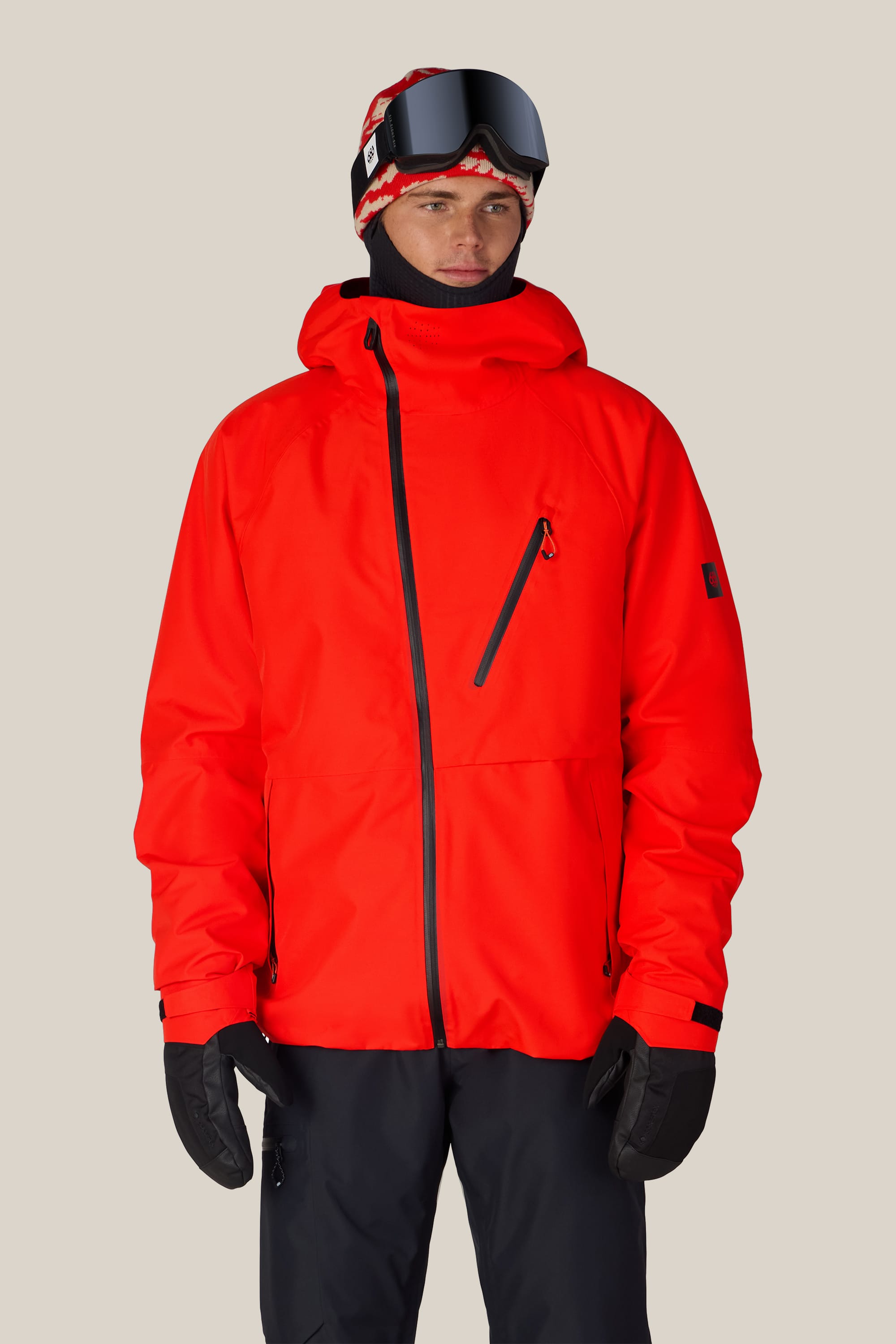 A person wearing the 686 Men’s Hydra™ Thermagraph® Jacket in bright red by 686, paired with black gloves, black pants, a red and black ski hat, and ski goggles, standing against a plain light background.