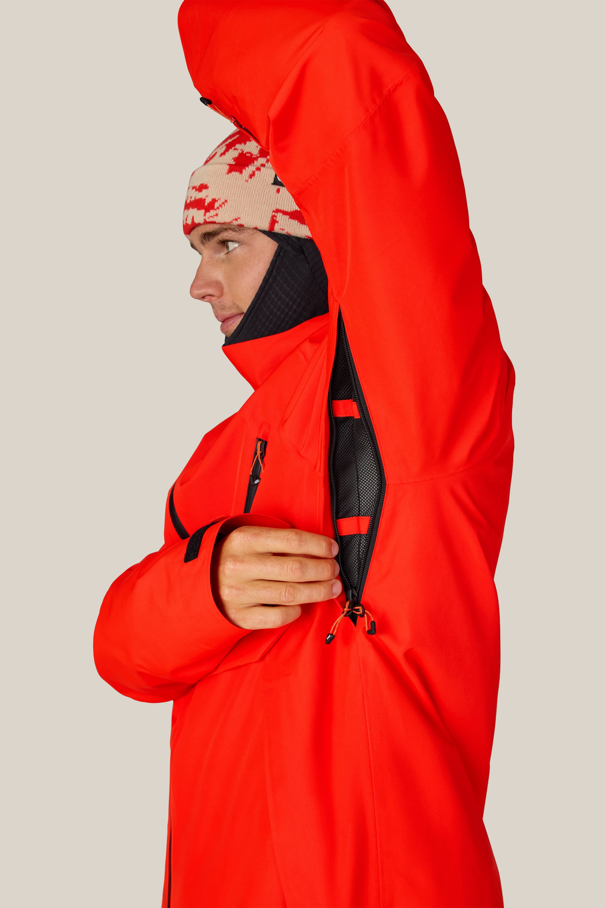 Wearing the 686 Men’s Hydra™ Thermagraph® Jacket, a person in a bright red style and patterned beanie lifts their arm to open the underarm zippered vent, revealing the black mesh lining inside this 686 jacket.