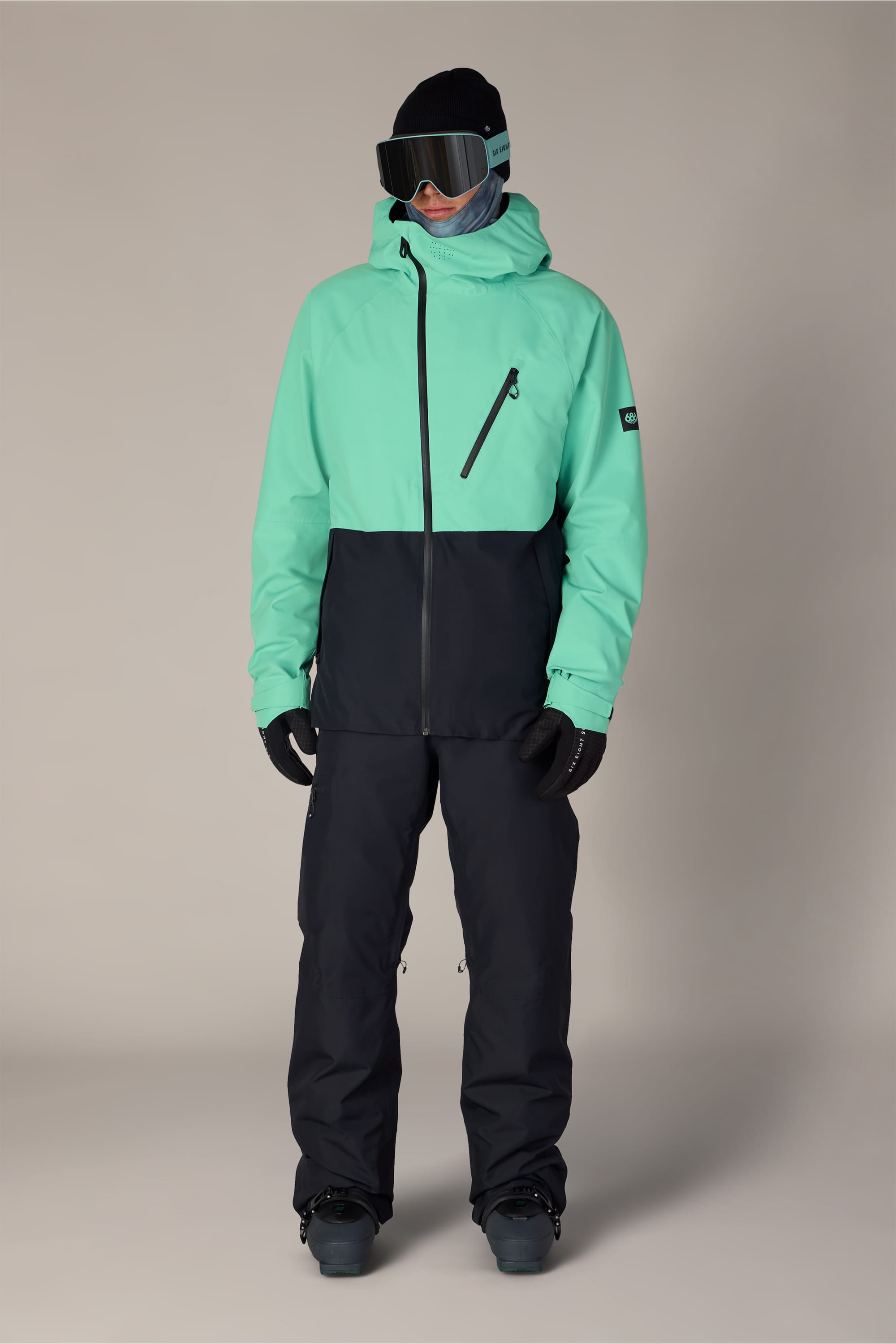 A person wearing the 686 Men’s Hydra™ Thermagraph® Jacket in mint green and black by 686, paired with black ski pants, gloves, ski goggles, and a beanie, stands against a plain light background. Their face is fully covered by the hood of the jacket, goggles, and a neck gaiter.