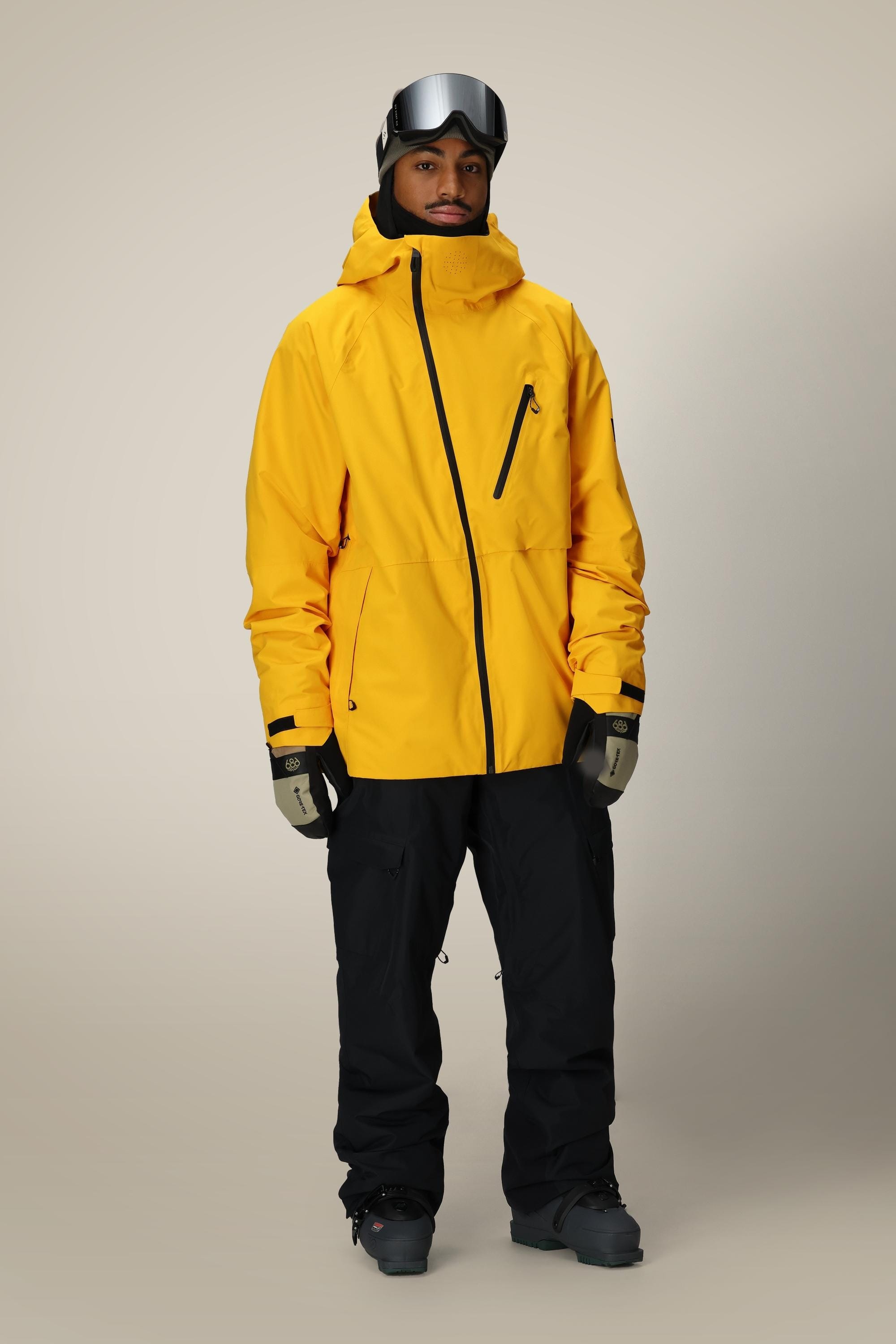 A person wears the 686 Men's Hydra™ Thermagraph® Jacket in yellow, paired with black snow pants, black gloves, gray ski goggles on their head, and black boots, standing against a plain light background.