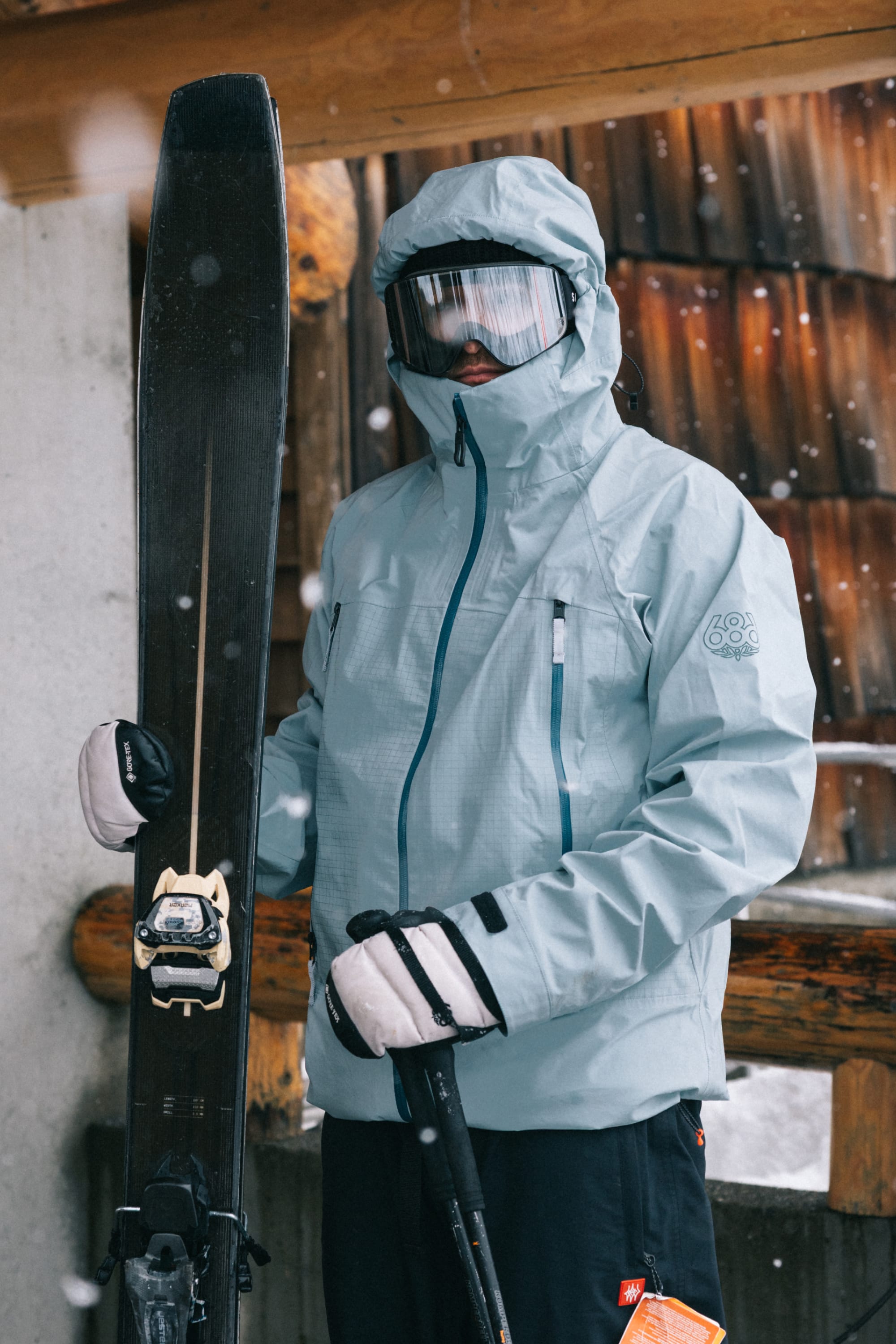 A person wearing the 686 Men’s SMARTY® 3-In-1 Rodeo™ 3L Jacket by 686, along with gloves and goggles, holds a pair of skis and ski poles while standing outdoors near a wooden building as snow falls around them.