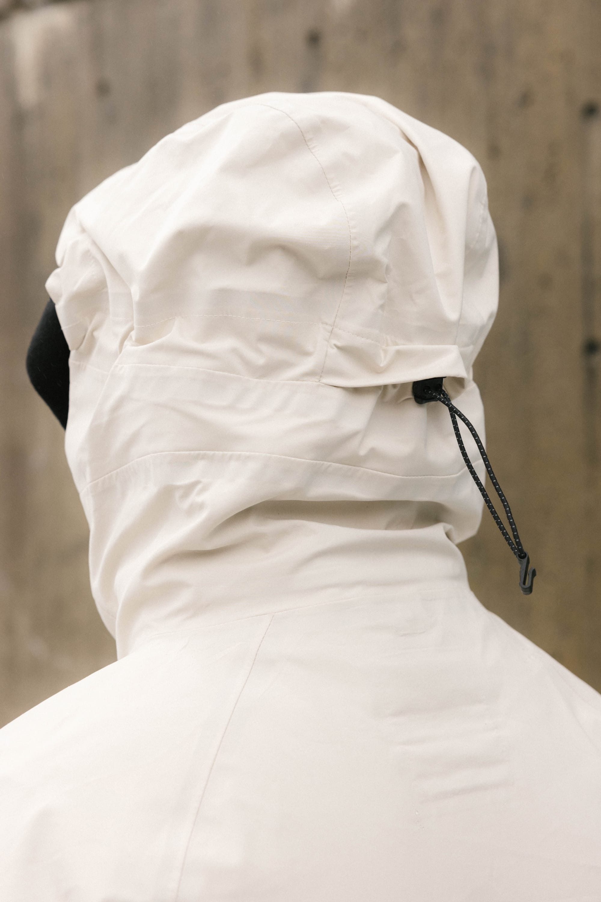 A person in a 686 Men's SMARTY® 3-In-1 Rodeo™ 3L Jacket with a black adjustable drawstring stands before a textured concrete wall. The shot, taken from behind, showcases the hood and upper back of the stylish 686 jacket.