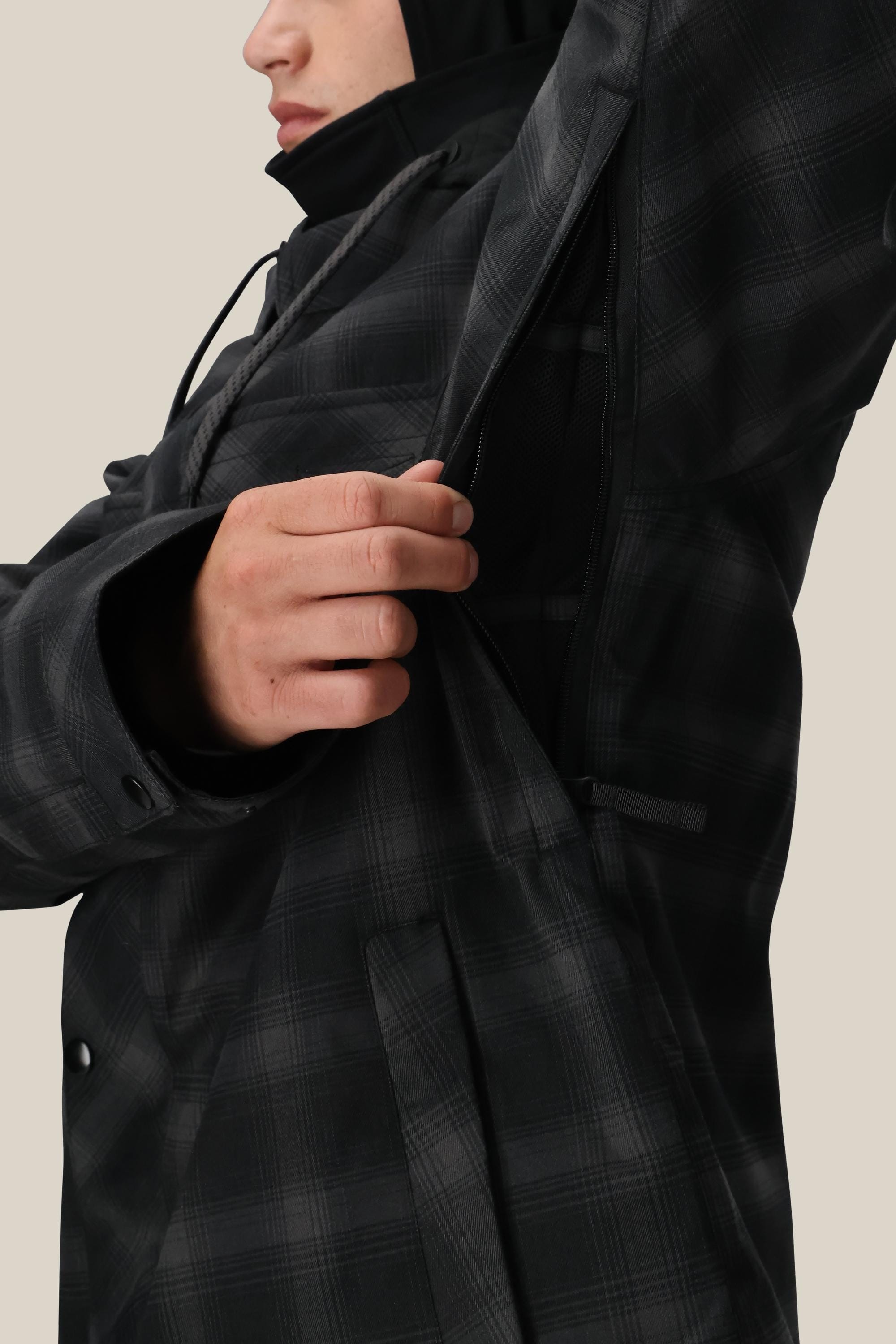 A person wearing the 686 Men's Woodland™ Insulated Jacket with a black plaid pattern unzips an underarm vent, partially revealing their face and hand against a neutral background.