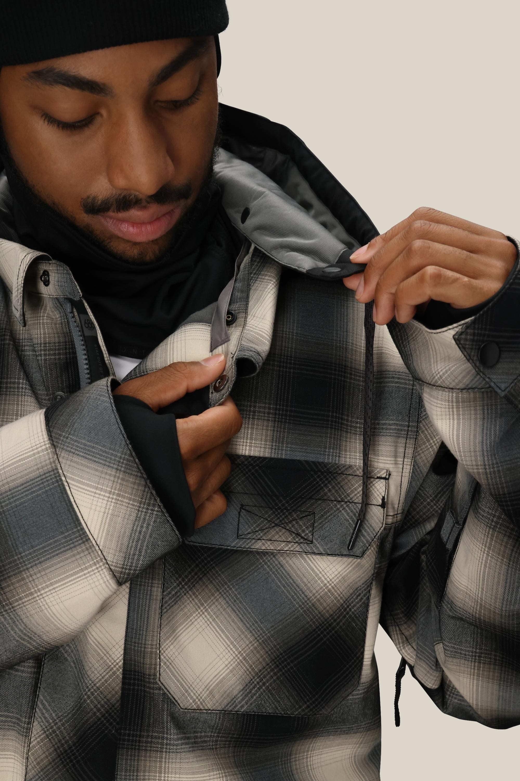 A person wearing a black beanie and the 686 Men’s Woodland™ Insulated Jacket by 686, featuring a black, gray, and white checkered pattern, adjusts the collar of their jacket while looking down.