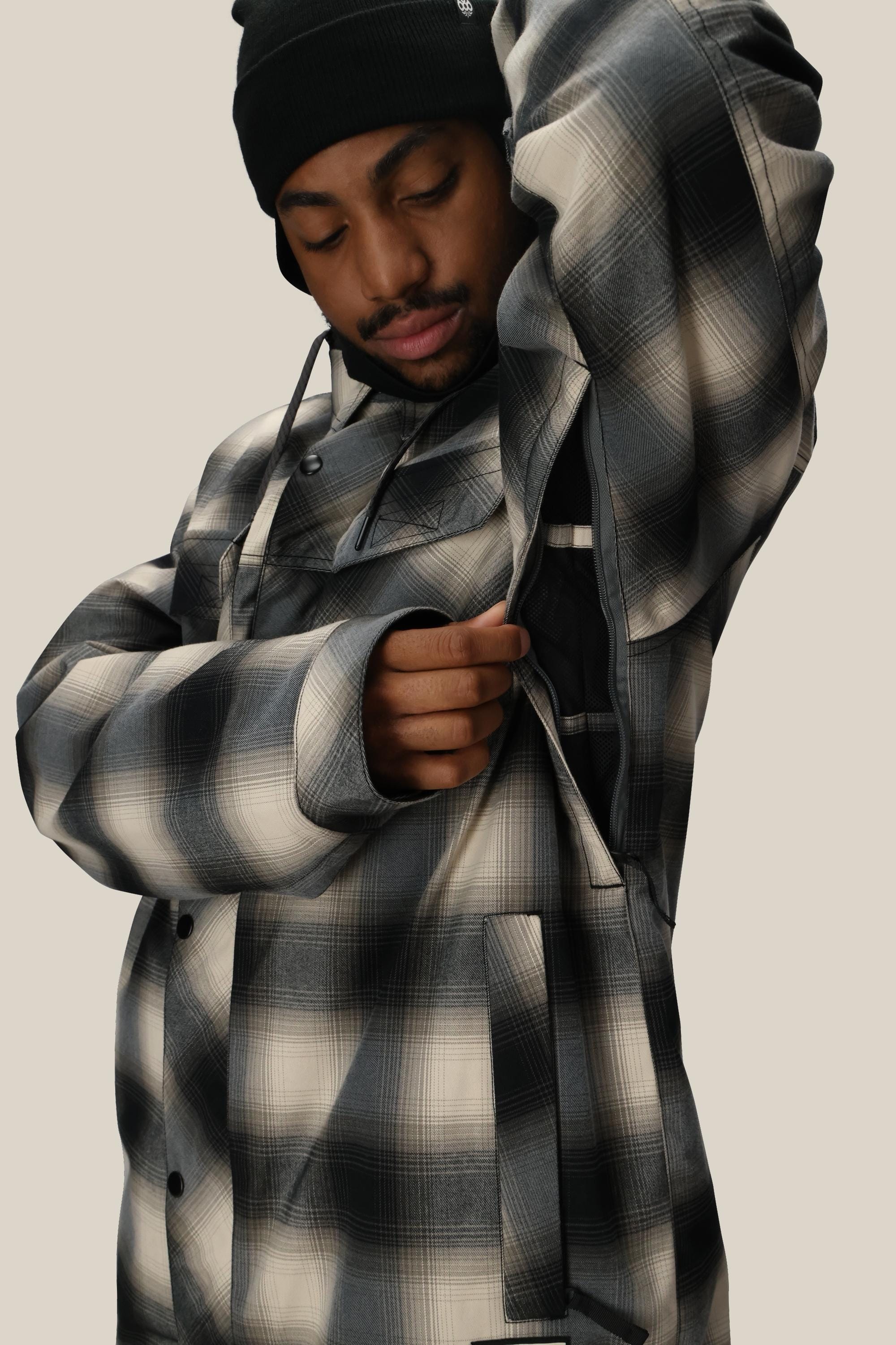 A person in a black beanie adjusts the 686 Men's Woodland™ Insulated Jacket, opening a zippered pocket under their arm and looking down, all against a plain background.