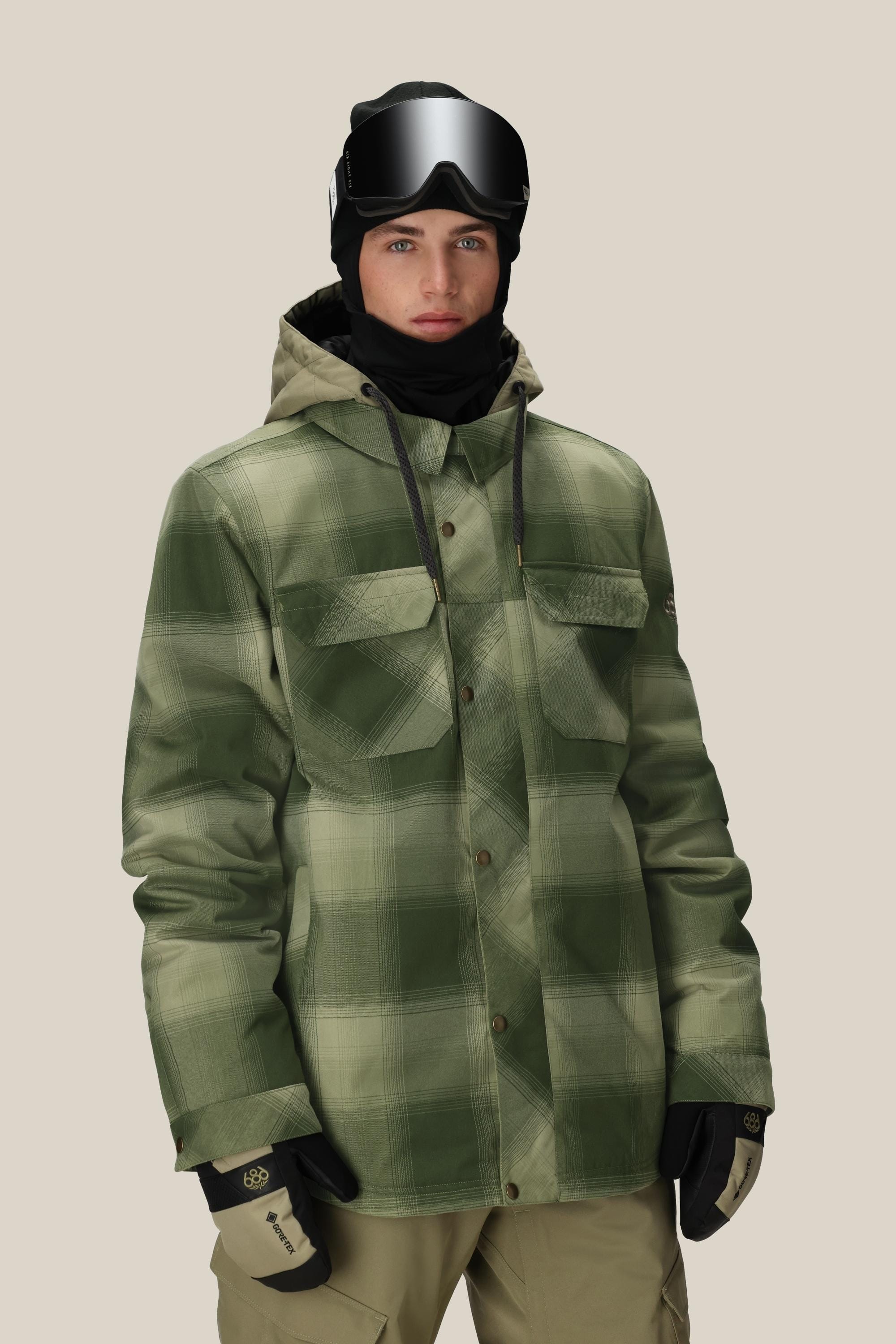 A person in a 686 Men's Woodland™ Insulated Jacket stands against a plain beige background, wearing tan snow pants, black gloves, ski goggles, and a hood.