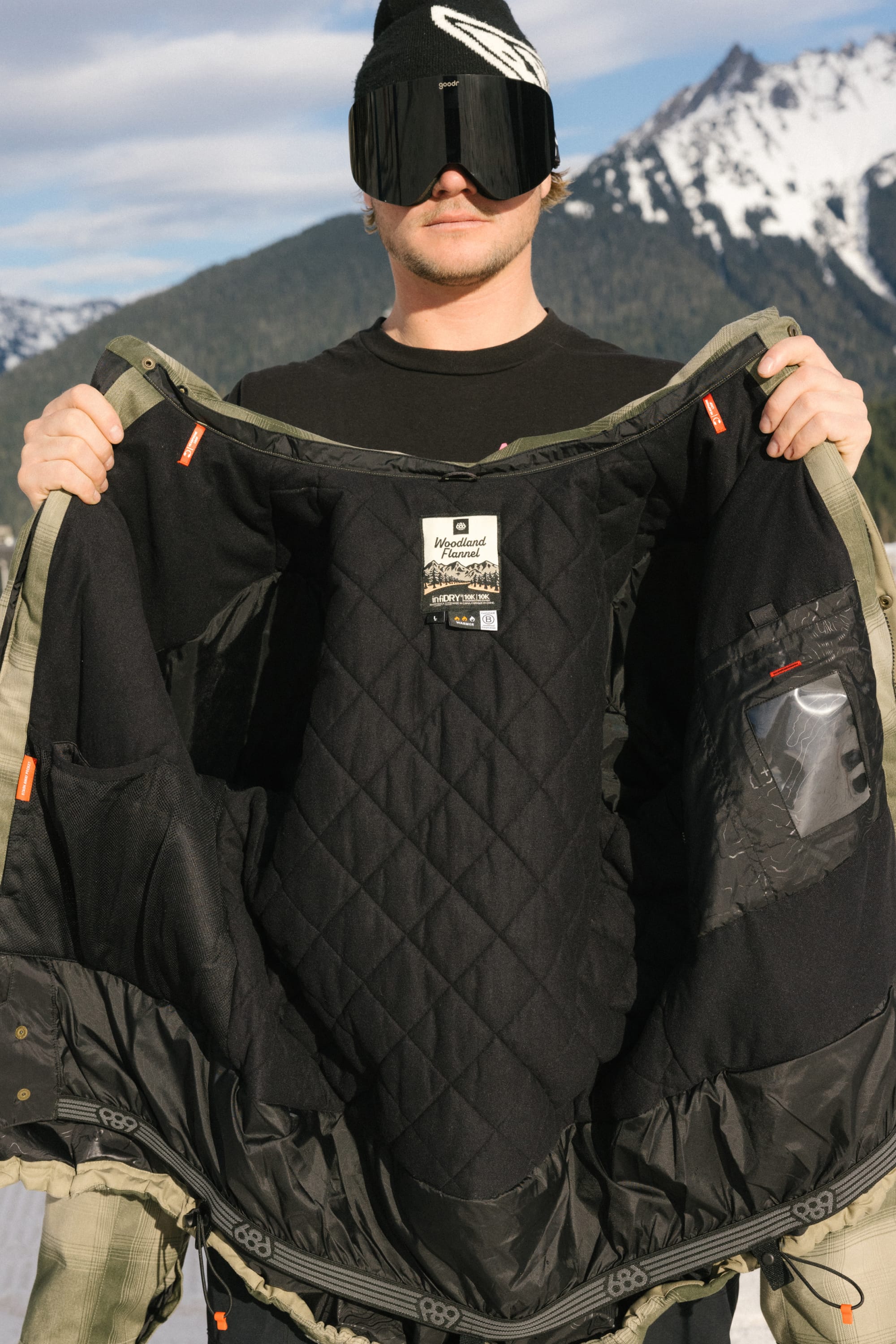 Wearing ski goggles and a beanie, a person holds open the 686 Men's Woodland™ Insulated Jacket, showing its quilted black interior with snowy mountains in the background.