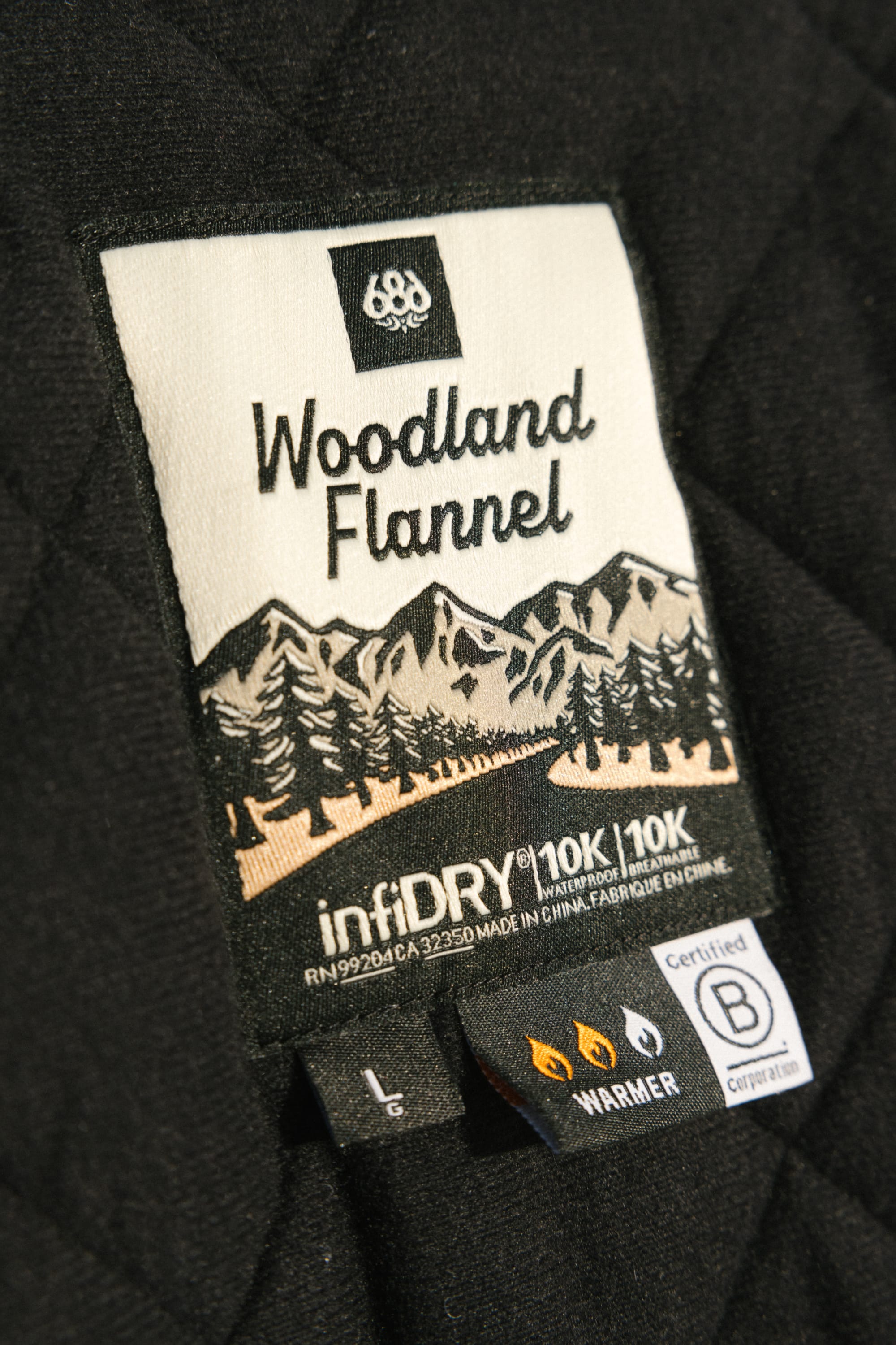A close-up of the label on a black quilted 686 Men's Woodland™ Insulated Jacket shows "Woodland Flannel" with mountain and tree graphics, plus infiDRY 10K/10K, ‘warmer’, and B certified details.