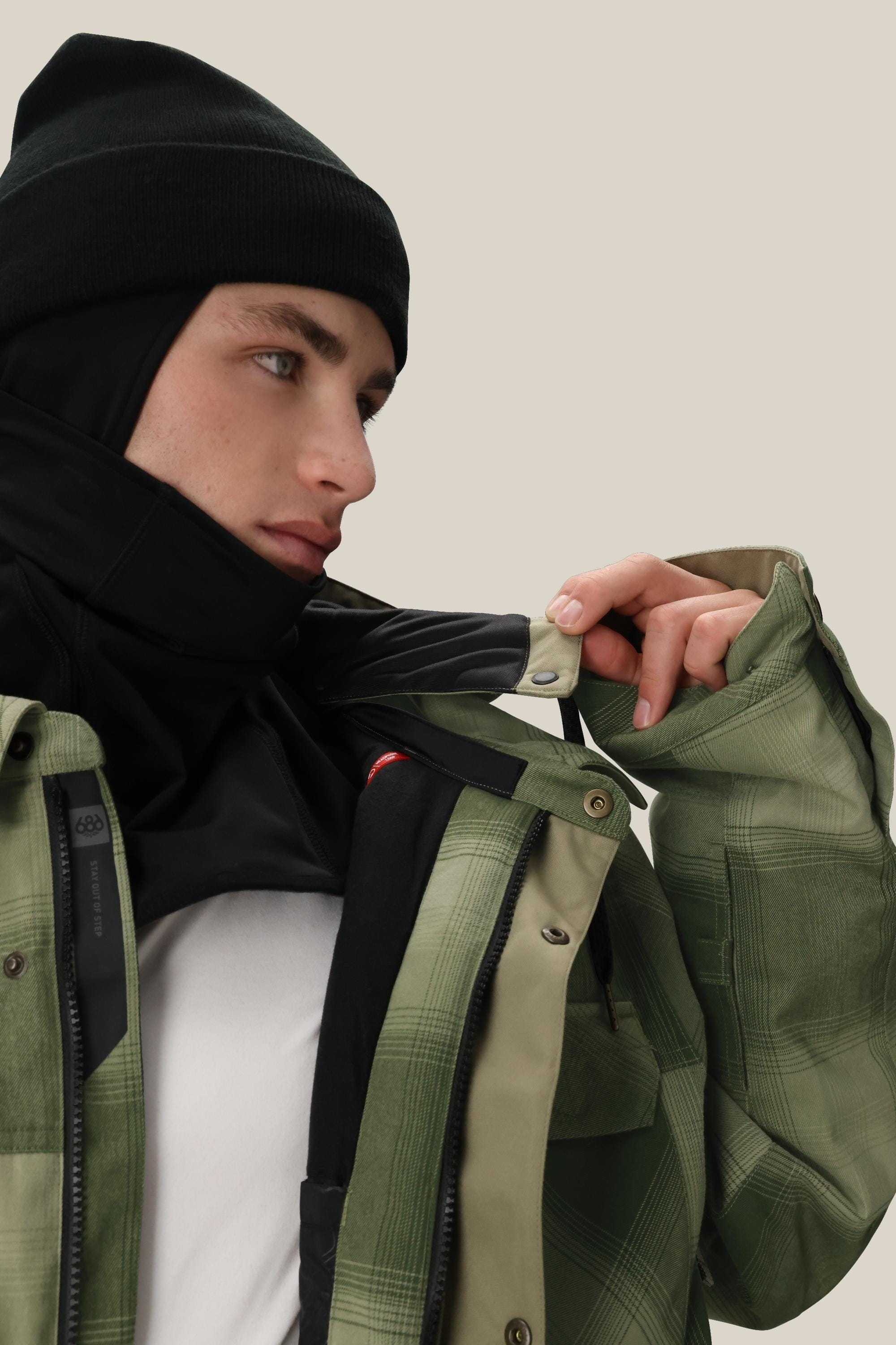 A person wearing a black beanie, black neck gaiter, and white shirt adjusts the collar of their 686 Men's Woodland™ Insulated Jacket by 686 against a plain beige background.