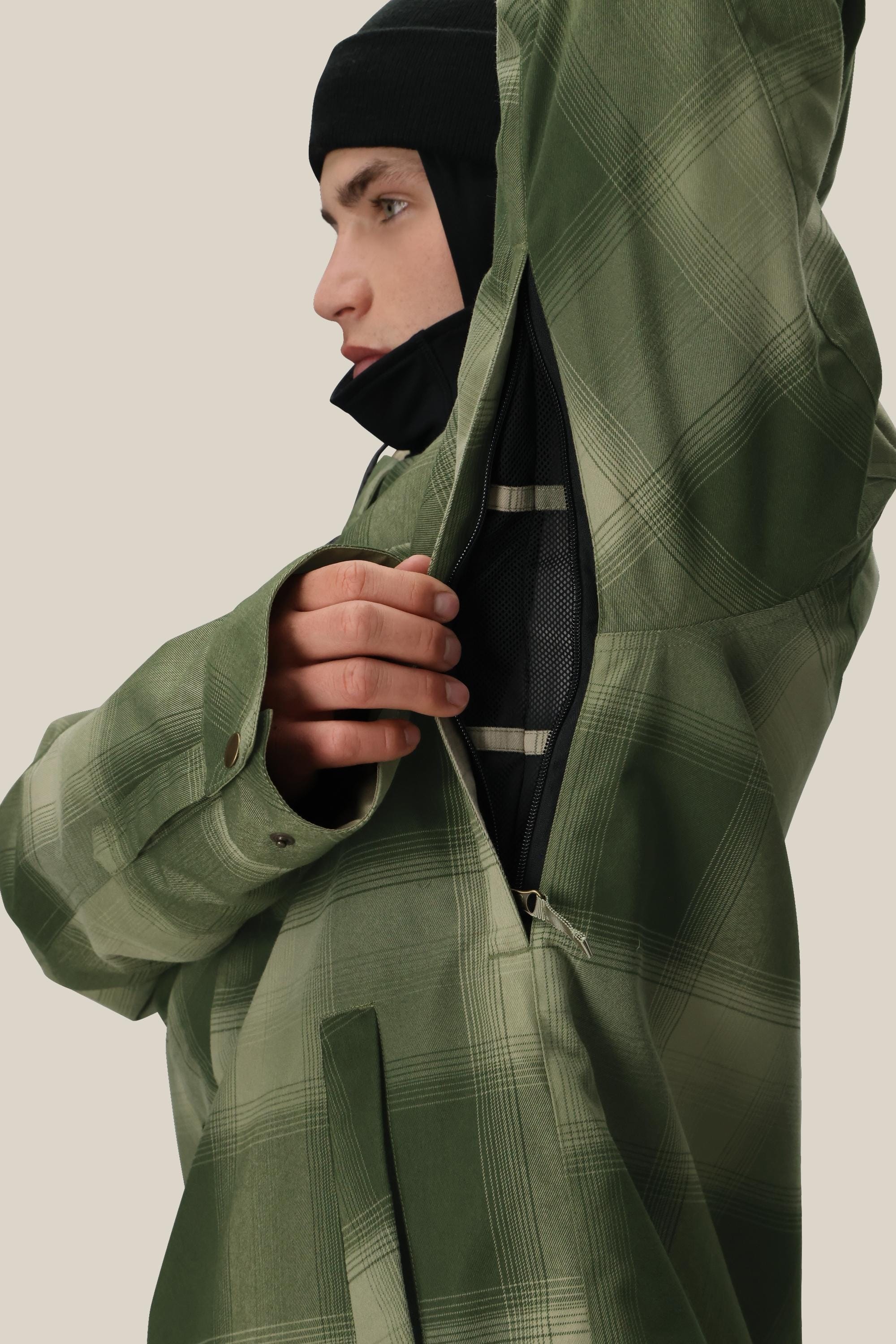 A person wearing the 686 Men's Woodland™ Insulated Jacket in green plaid with a black beanie unzips an underarm vent to reveal mesh lining. Shown from the side against a neutral background.
