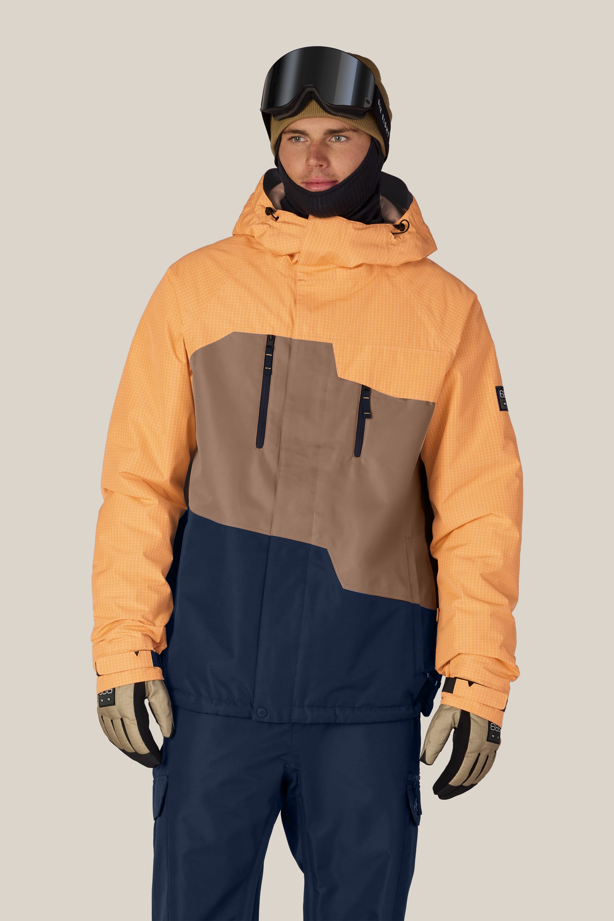 A person wearing the 686 Men’s Geo™ Insulated Jacket in peach, brown, and navy, along with ski pants, gloves, goggles, and a beanie stands against a plain background.