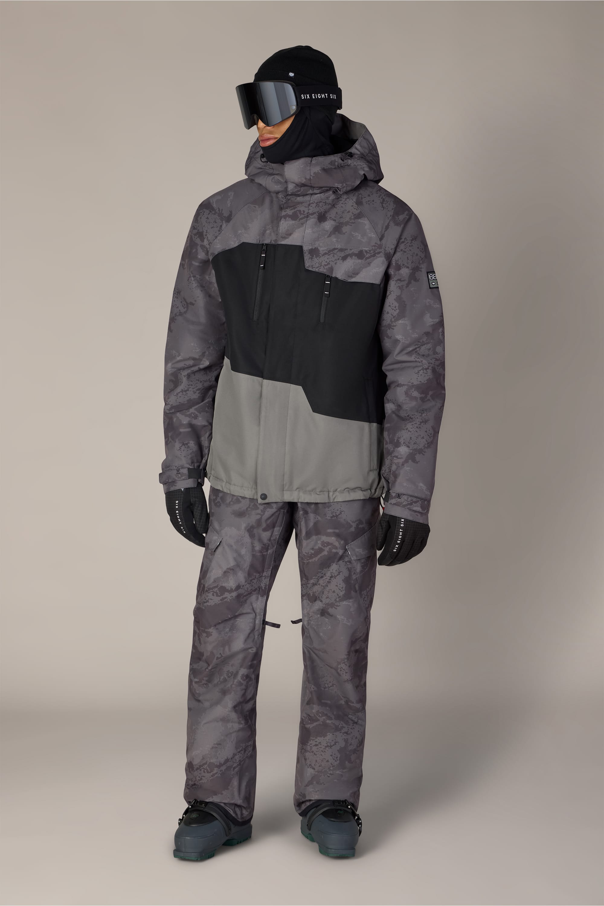 Wearing the 686 Men’s Geo™ Insulated Jacket in a gray and black camouflage pattern, along with matching pants, black gloves, winter boots, ski goggles, and a black face covering, a person stands against a plain light background.