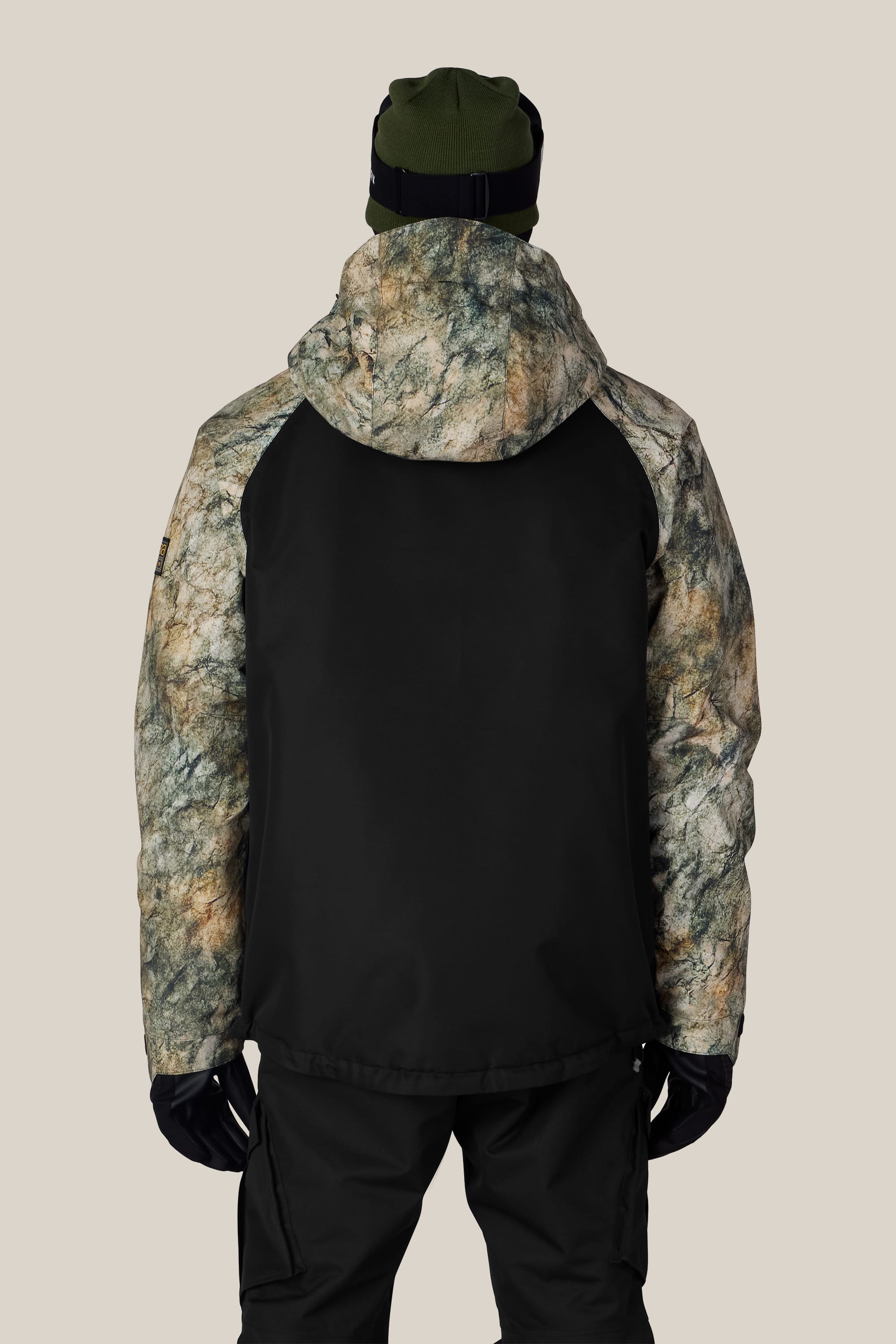 A person wearing the 686 Men’s Geo™ Insulated Jacket in black and camouflage by 686, along with black pants, black gloves, and a green beanie, stands with their back to the camera against a plain light background.