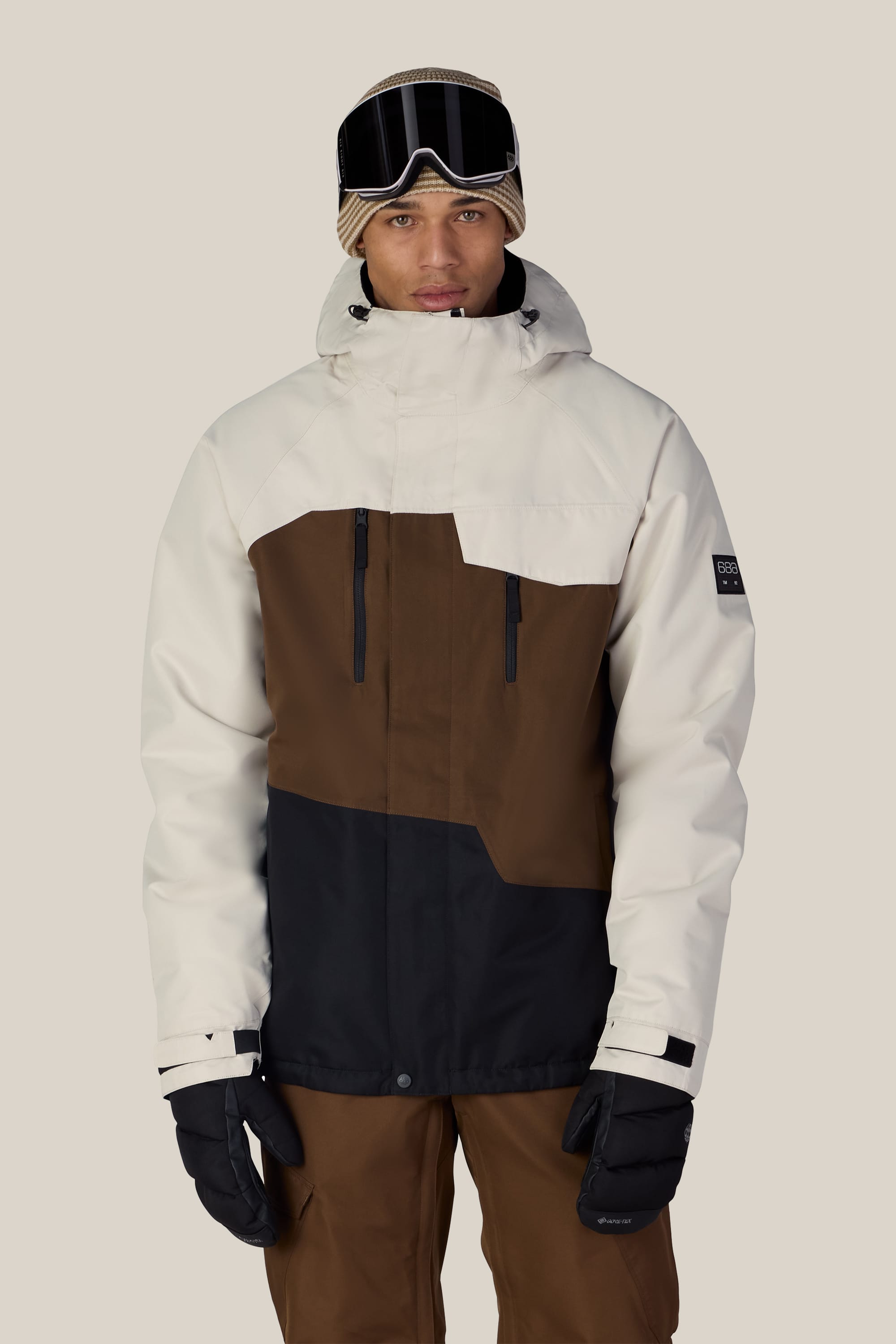 A person wearing the 686 Men’s Geo™ Insulated Jacket by 686—a woven, color-blocked ski jacket in white, brown, and black—paired with matching brown pants, black gloves, ski goggles, and a beige knit hat stands facing forward against a plain background.