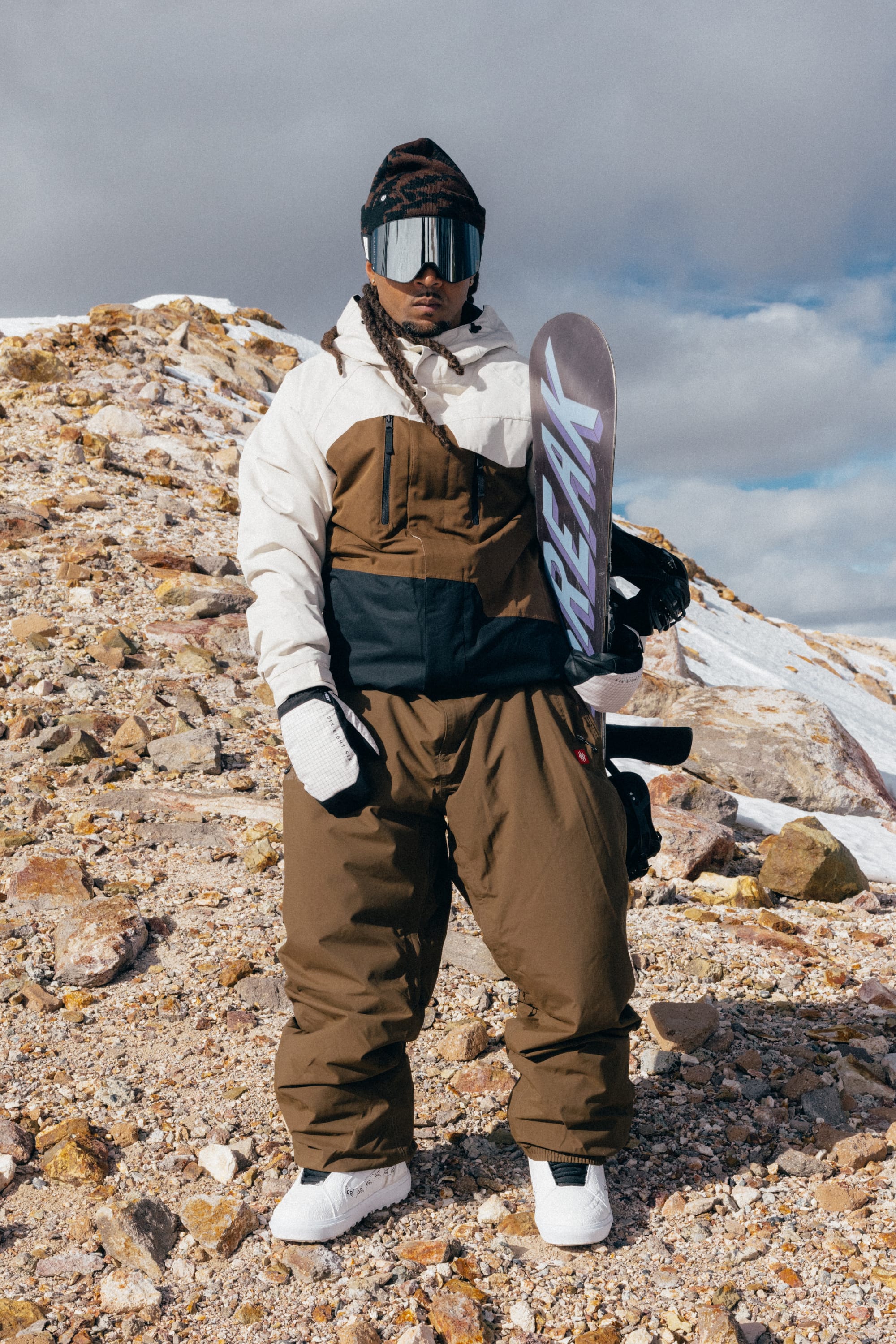 Wearing the 686 Men’s Geo™ Insulated Jacket, a person in brown and white snow gear stands on a rocky, snowy mountain slope holding a snowboard. They are also equipped with gloves, a helmet, and reflective goggles.
