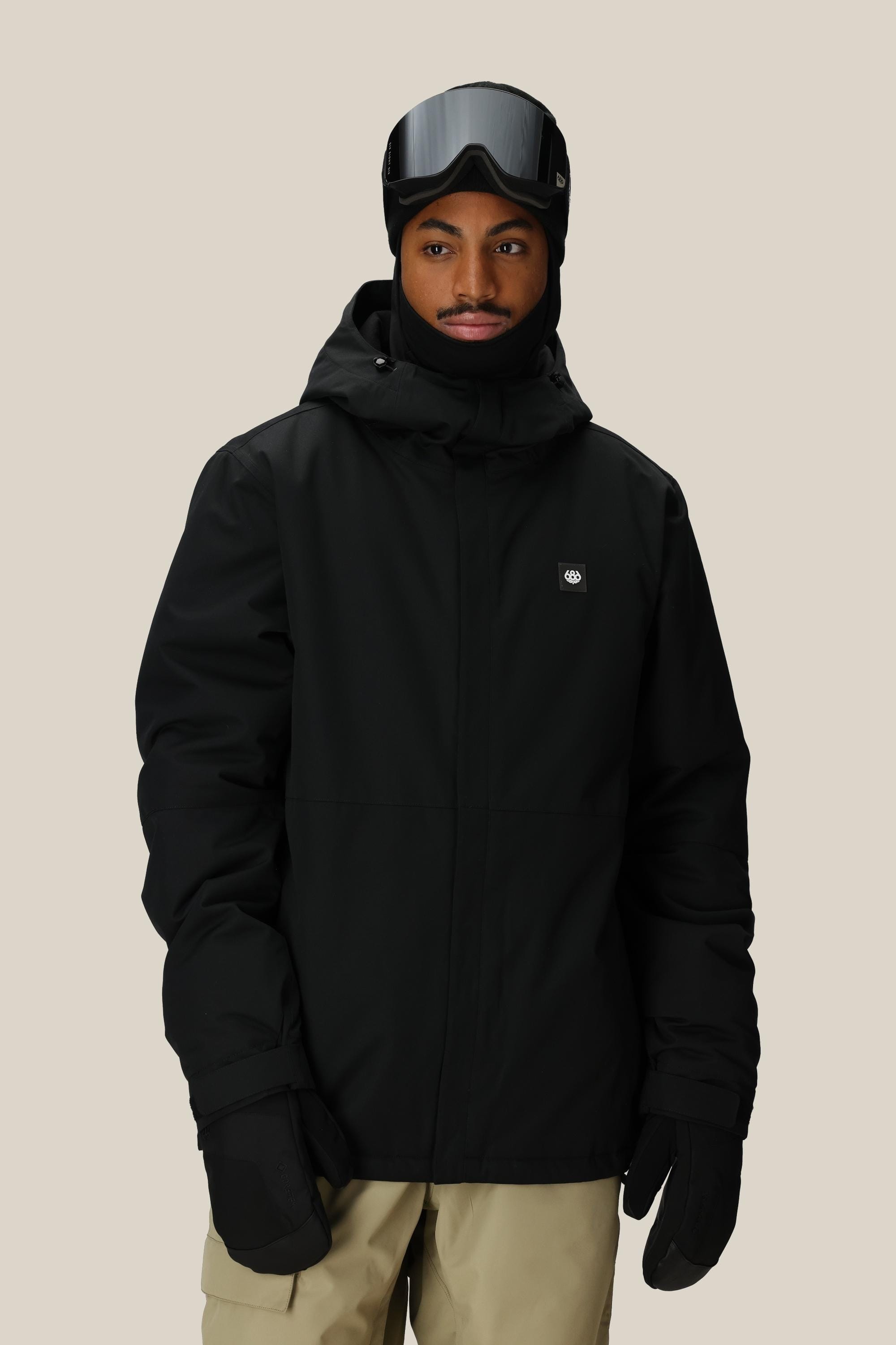 A person wearing the 686 Men’s Foundation™ Insulated Jacket by 686, paired with ski goggles and gloves, stands against a plain background. They are also dressed in light brown pants and have a black face covering beneath the jacket’s hood.