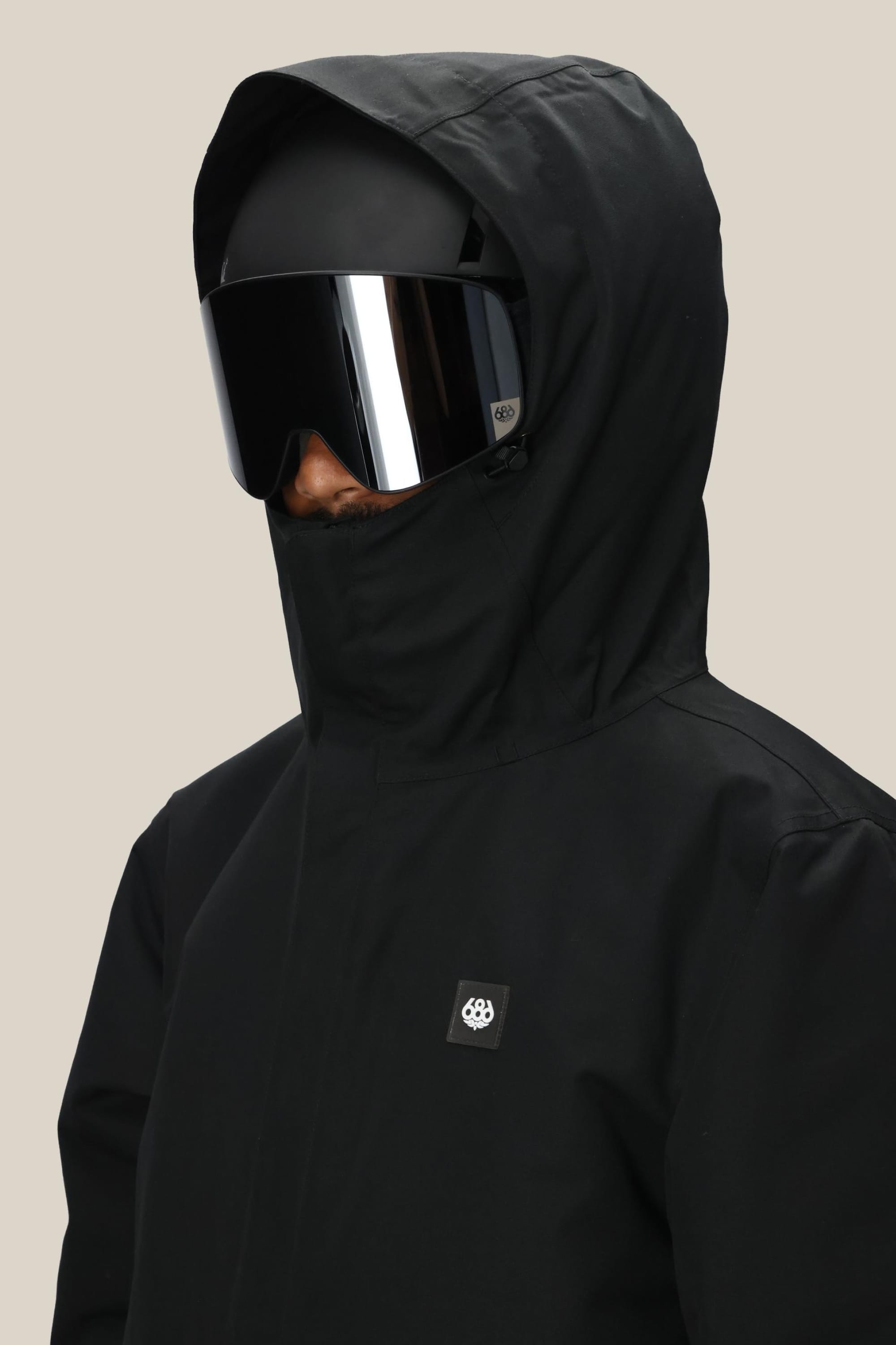 A person wearing the 686 Men's Foundation™ Insulated Jacket in black with a hood and subtle chest logo, paired with large reflective ski goggles, stands slightly turned against a plain beige background.