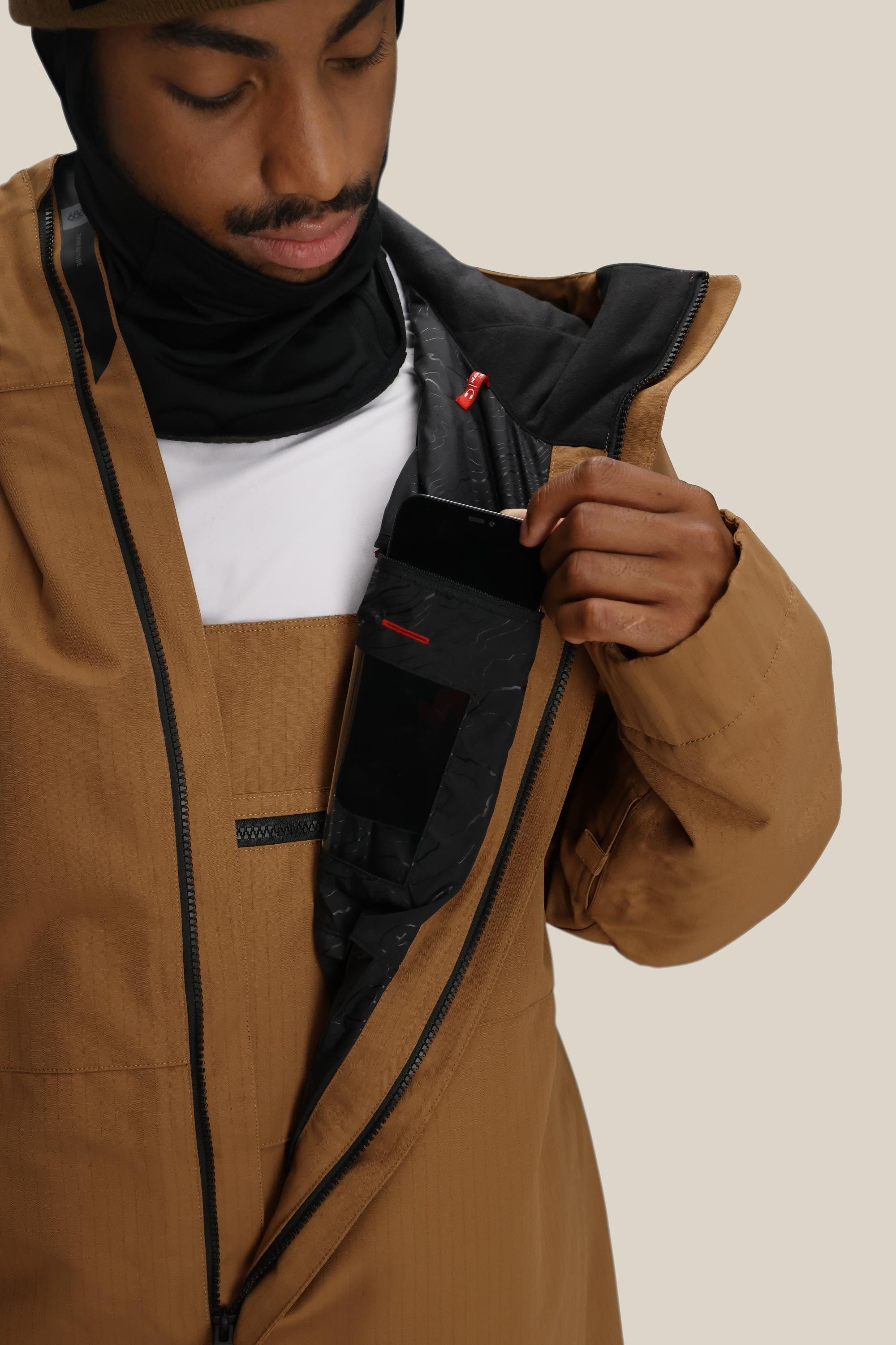 A person wearing a 686 Men's Foundation™ Insulated Jacket and a black head covering unzips the coat, revealing an inner chest pocket where they place a black smartphone.