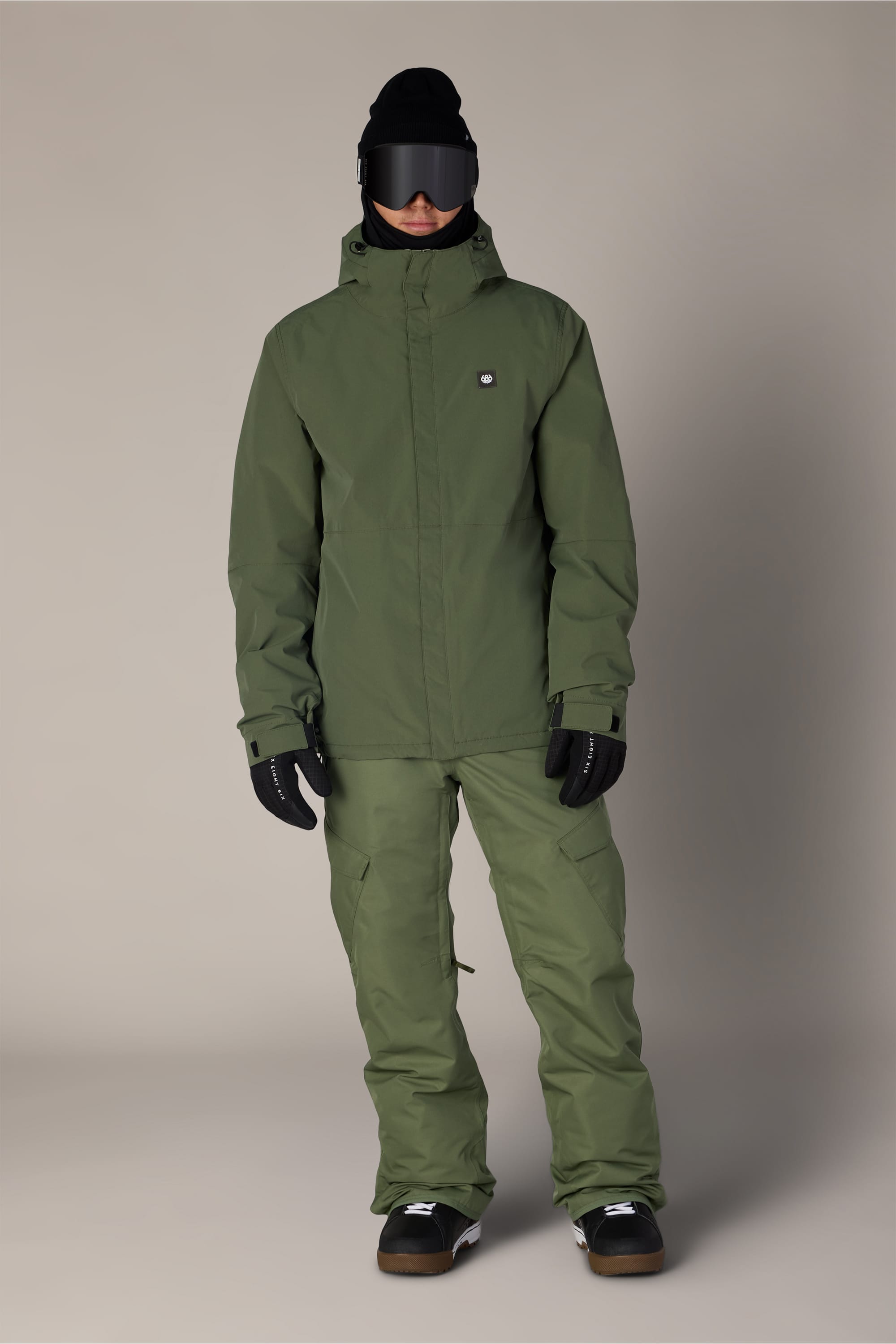 A person stands against a plain background, wearing the 686 Men’s Foundation™ Insulated Jacket in olive green along with matching pants, black gloves, black boots, a black beanie, and dark ski goggles.