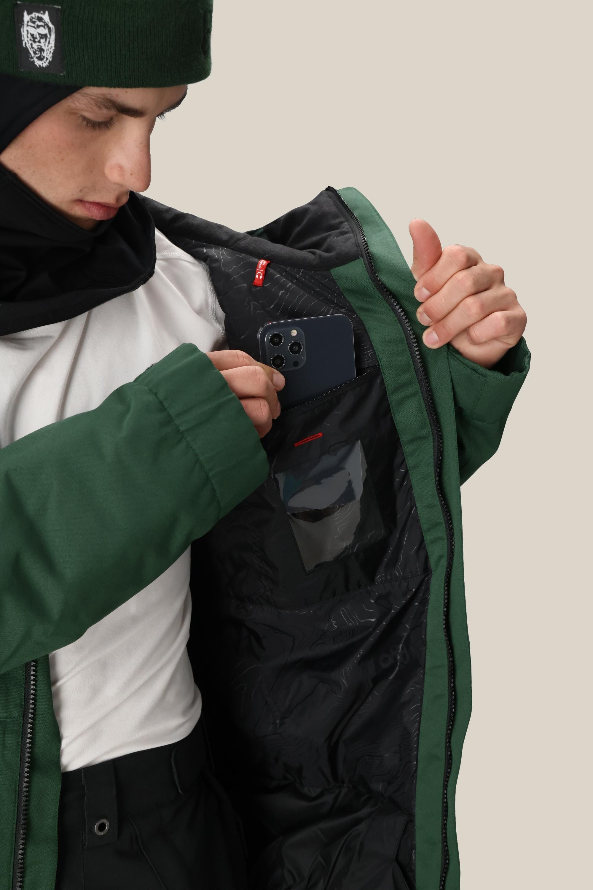 A man in a 686 Men's Foundation™ Insulated Jacket and beanie shows the black interior pocket while placing his smartphone inside. The lining has a clear ID slot and red tag, highlighting the style of this 686 jacket.
