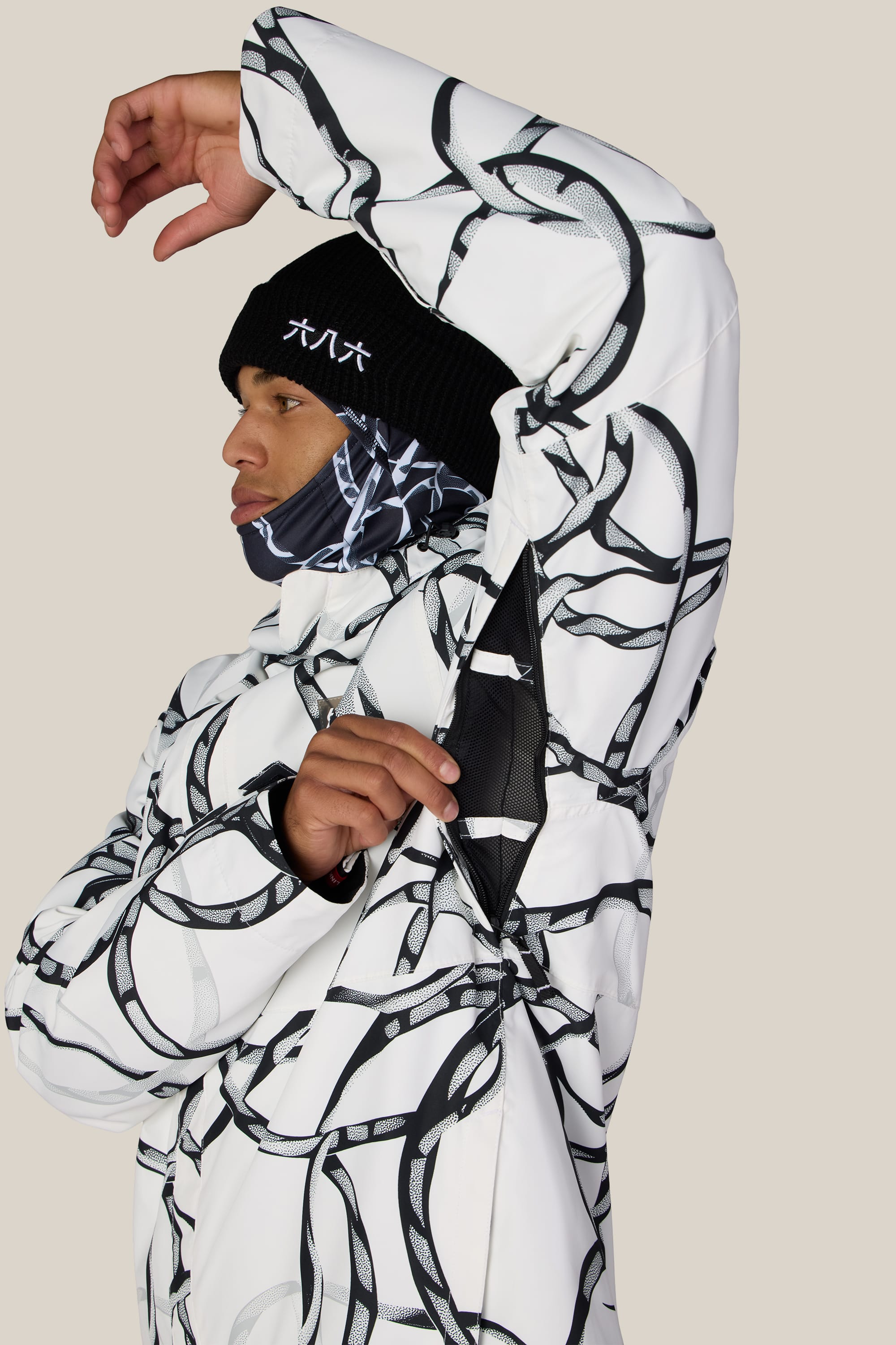 A person wearing the 686 Men’s Foundation™ Insulated Jacket in a white and black pattern opens the zippered underarm vent. They pair it with a black beanie featuring white text and a matching patterned face covering, set against a plain, light-colored background.