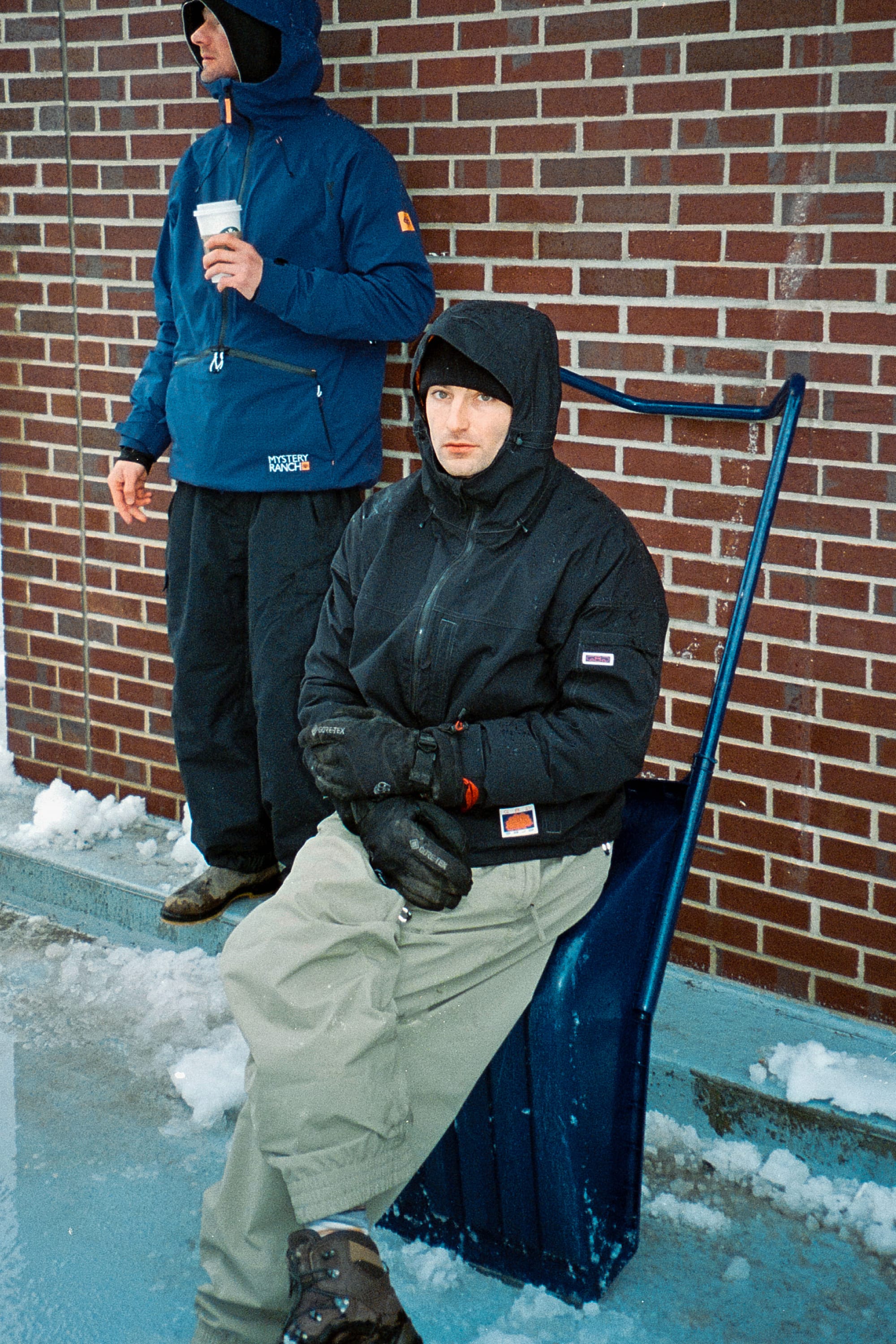 Two people are outside near a brick wall, dressed for winter. One, wearing the 686 Dojo® Jacket by 686, stands holding a cup while the other sits on a snow shovel. Snow and ice cover the ground around them.
