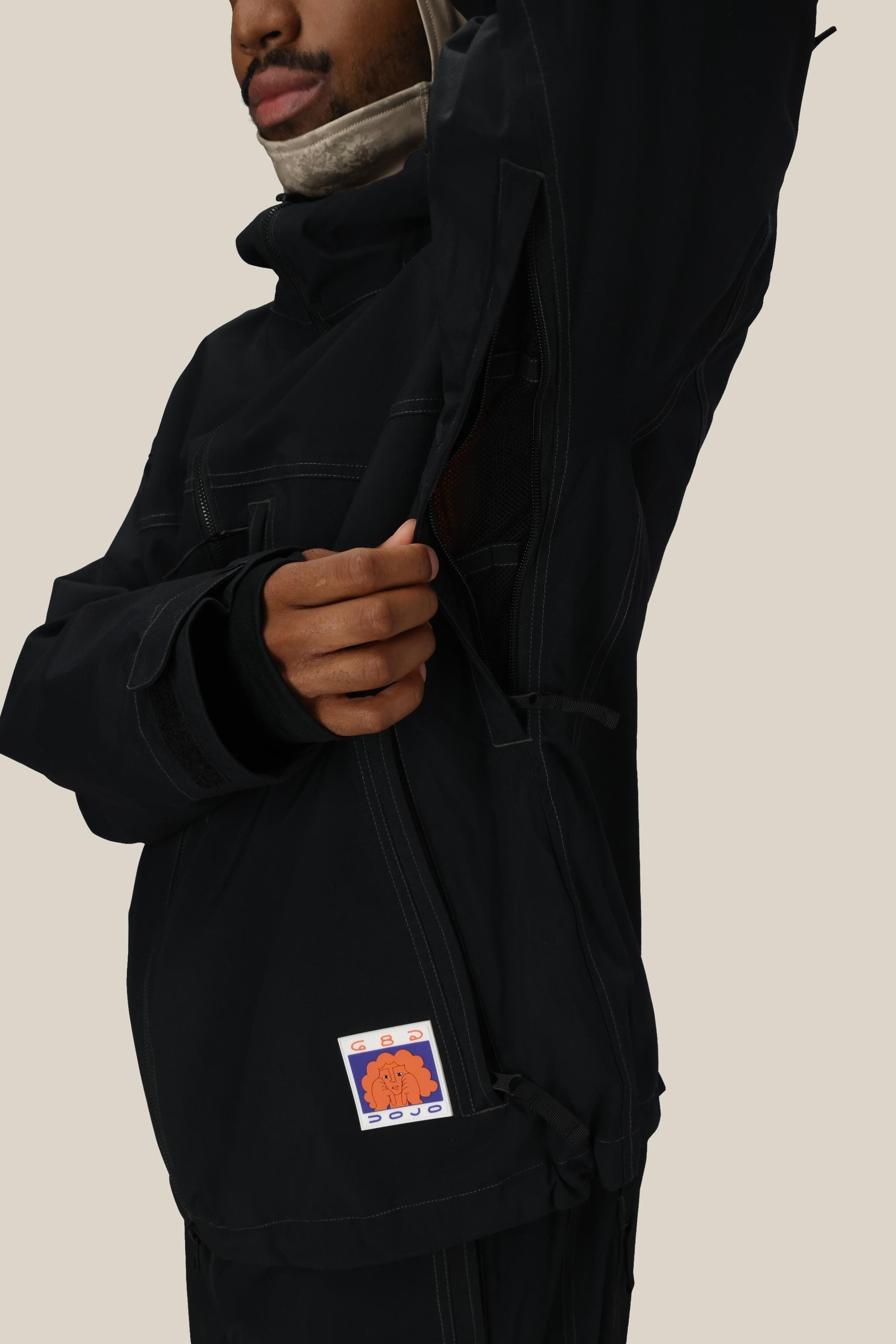 A person wears the 686 Dojo® Jacket by 686, featuring a zippered vent left partly open and a colorful patch near the hem. Their face is partly visible against a plain beige background.