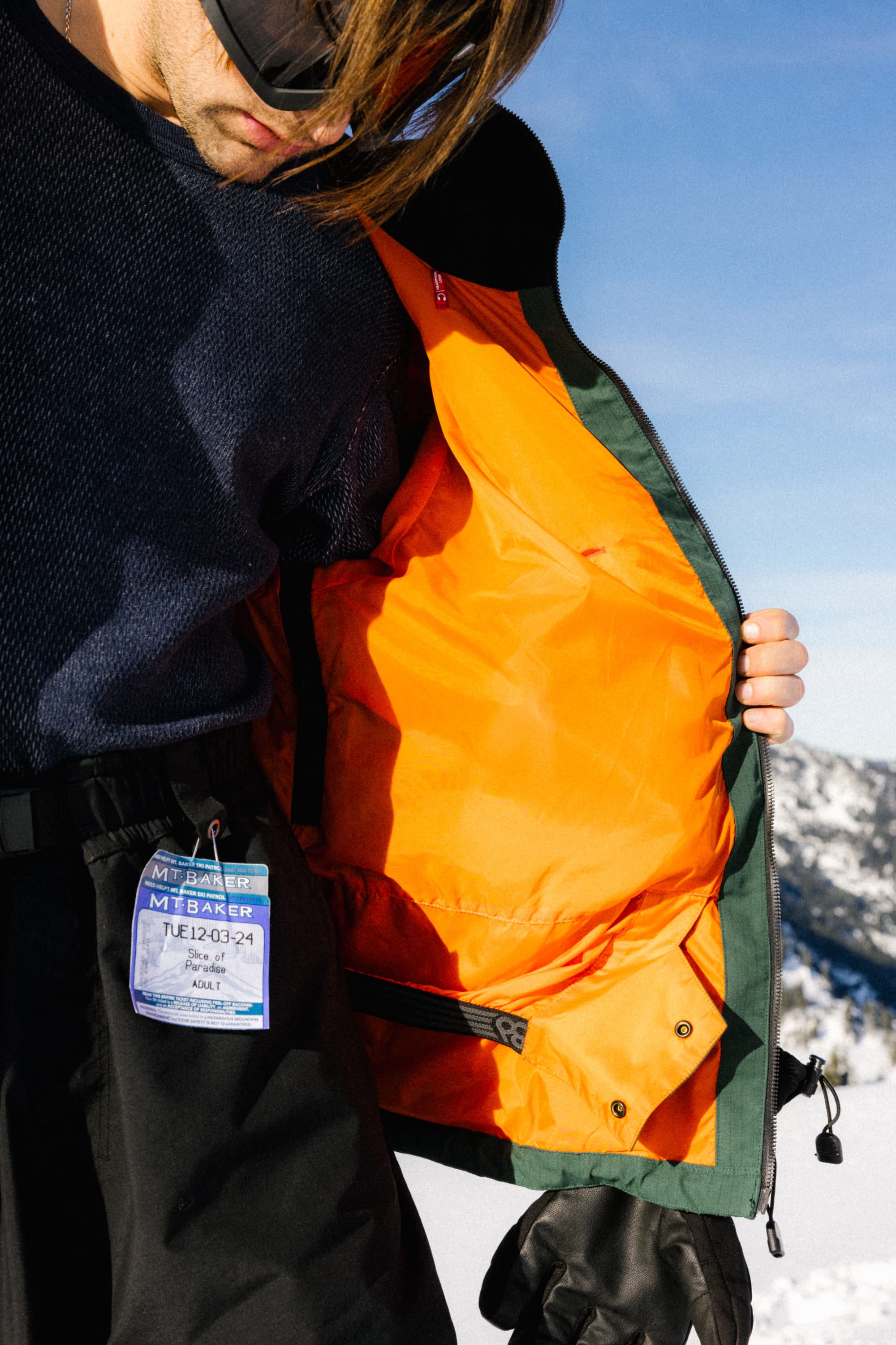 Wearing the 686 Dojo® Jacket with bright orange lining, a person shows its interior. A ski pass is clipped to their black snow pants, as they stand outdoors with snowy mountains and a blue sky behind them.