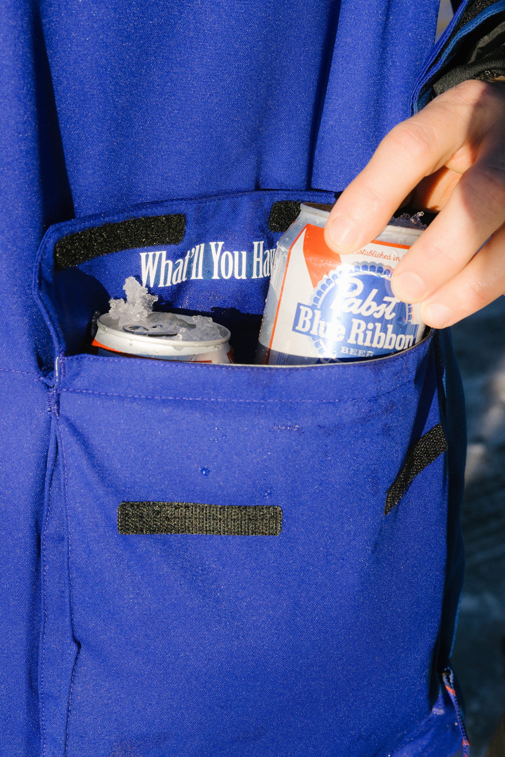 A hand places a can of Pabst Blue Ribbon into the pocket of the 686 Mens PBR Cooler Insulated Jacket by 686, which already holds another can and features What’ll You Have? above the pocket.
