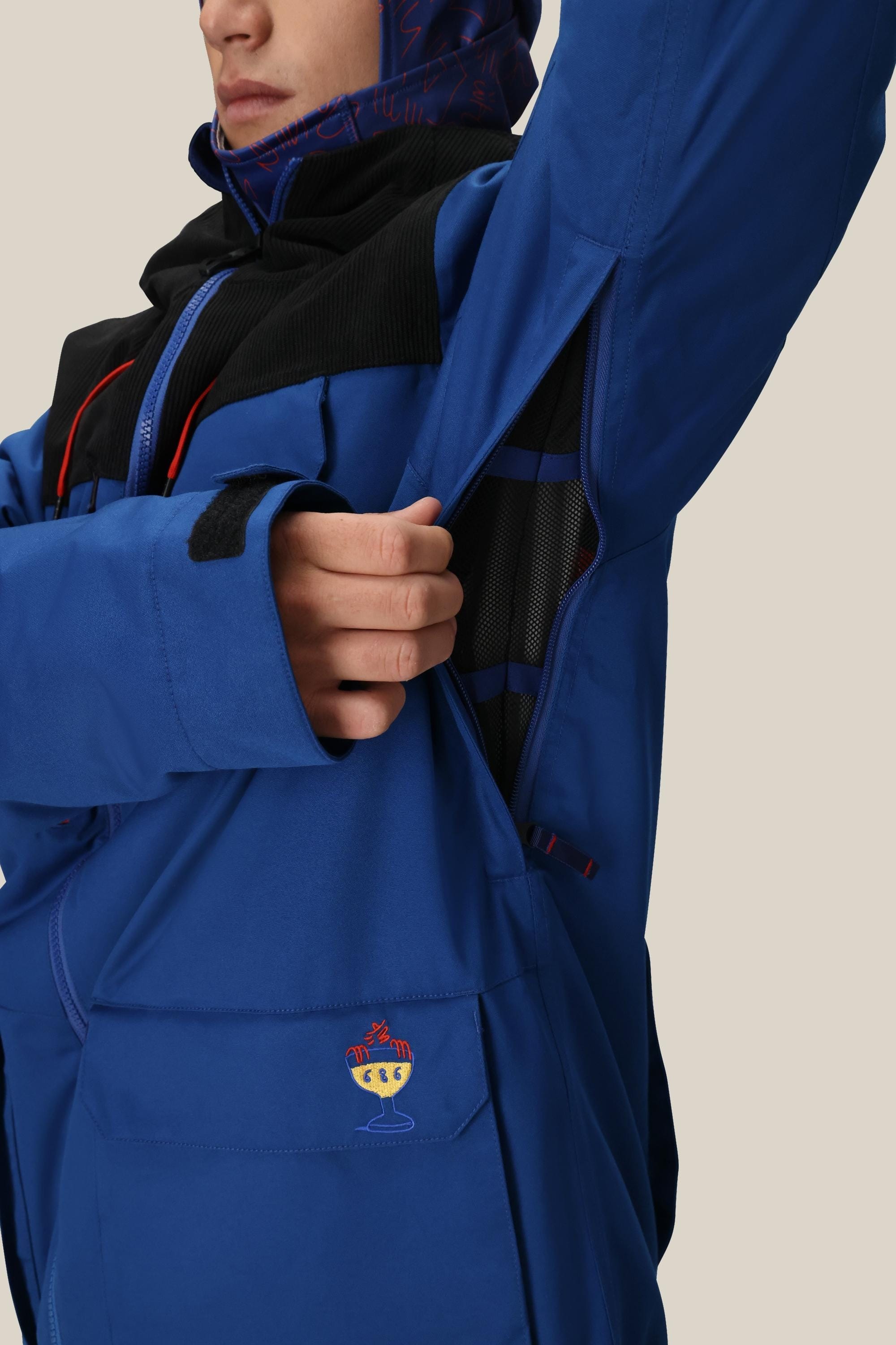 A person in the 686 Mens PBR Cooler Insulated Jacket, a blue waterproof woven polyester jacket with black accents and a cartoon cup patch on the lower sleeve, opens a zippered underarm vent. The face is partially visible.