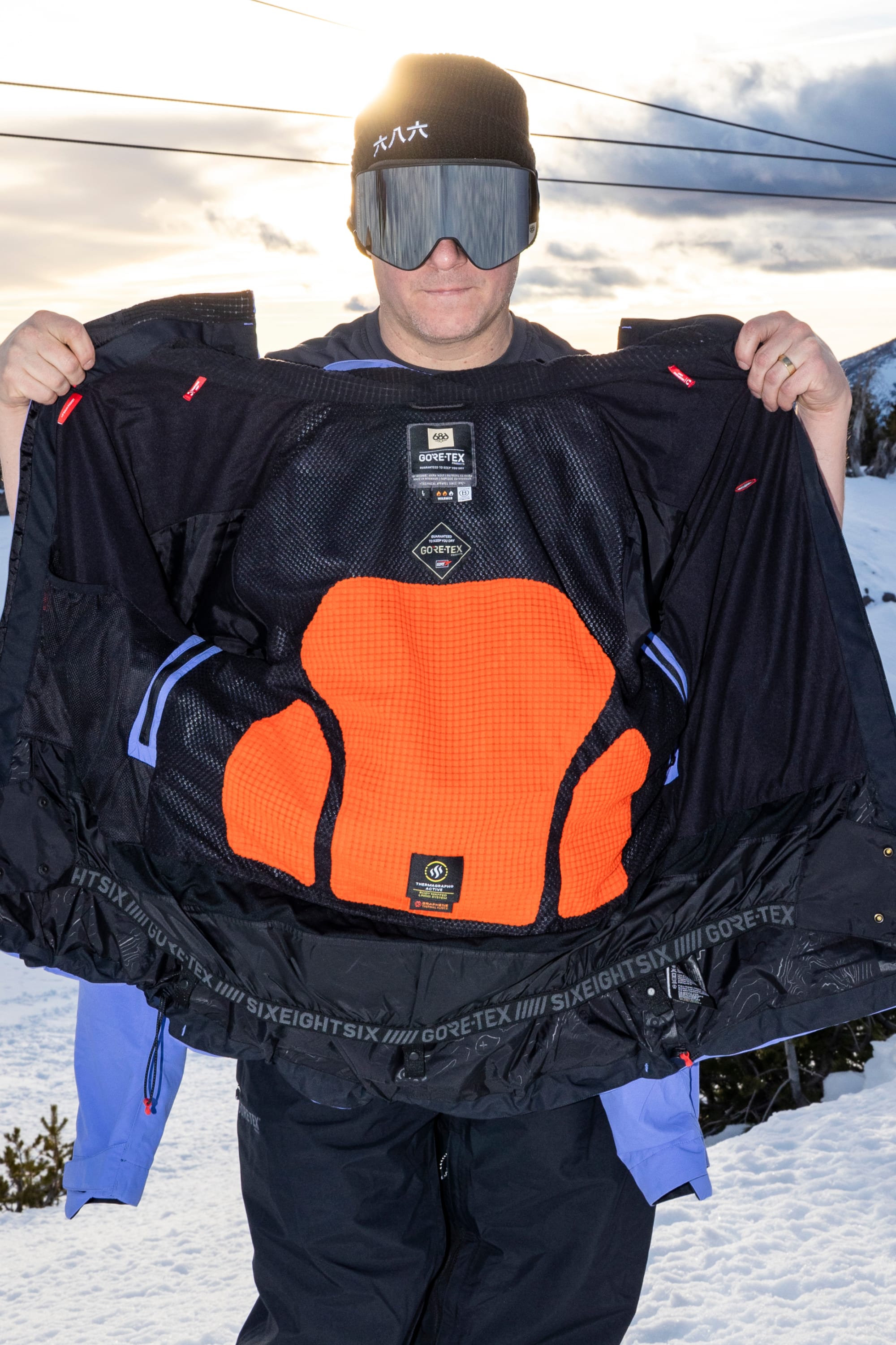 Dressed in the 686 Men’s GORE-TEX® GT Thermagraph® Jacket, a person stands outdoors in a snowy landscape with trees and a cloudy sky, opening their jacket to reveal the orange protective Thermagraph® insulation inside.
