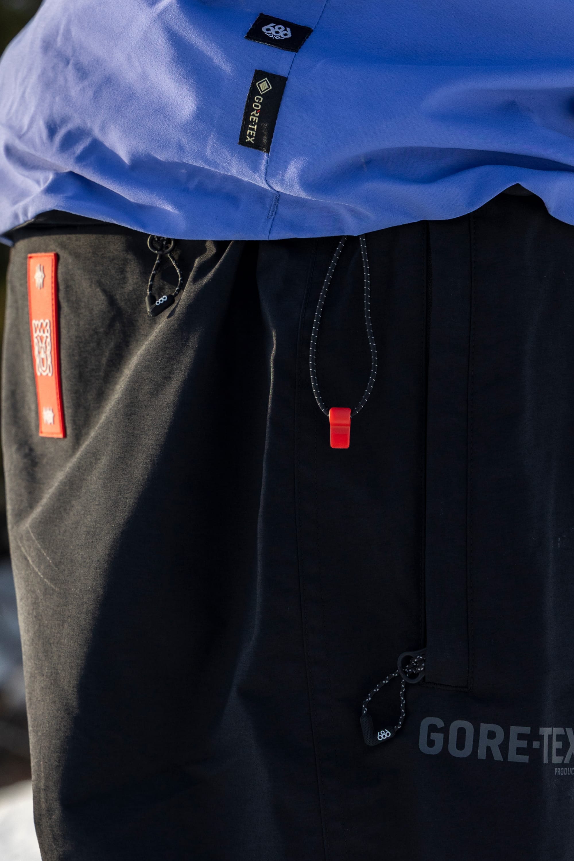 Close-up of a person wearing the 686 Men’s GORE-TEX® GT Thermagraph® Jacket in blue and black pants, featuring a red pull tab and visible white logo patches, outdoors in natural light.