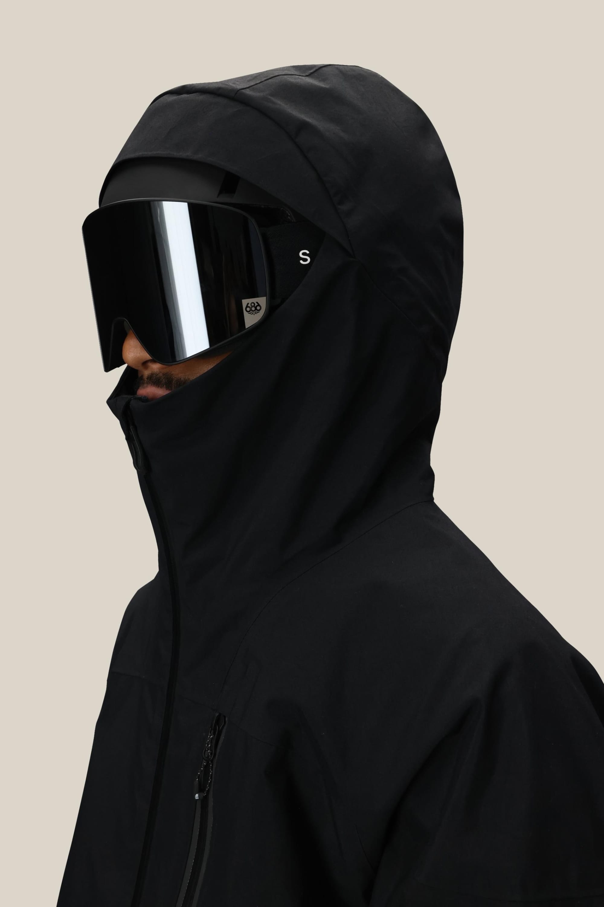 A man in a 686 Men's GORE-TEX GT Thermagraph® Jacket and large black reflective ski goggles is shown from the side against a plain beige background.