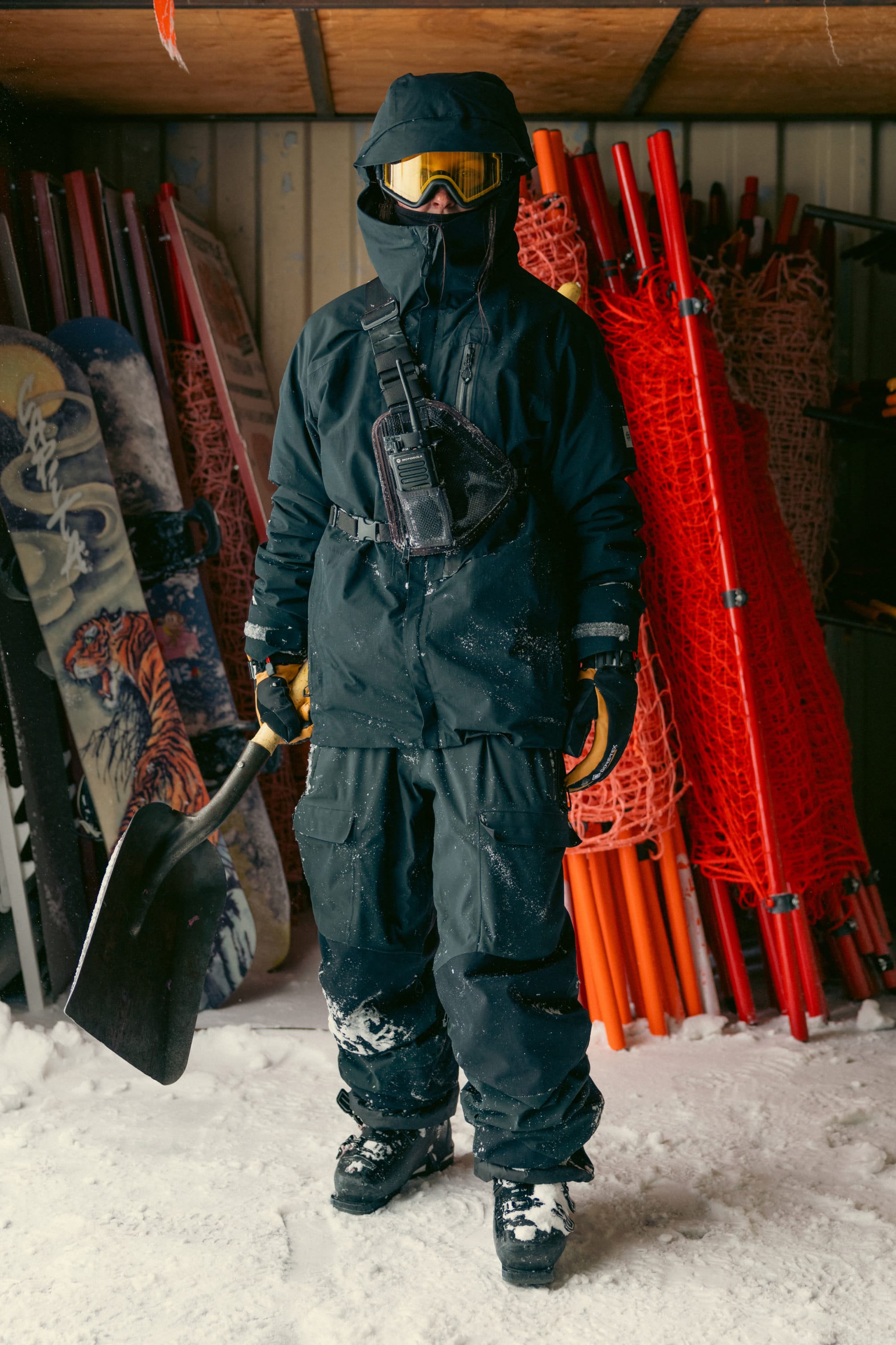 A person wearing the 686 Men’s GORE-TEX® GT Thermagraph® Jacket, along with black snow gear, goggles, and gloves, holds a snow shovel. Snowboards featuring tiger artwork and stacked orange safety nets are in the background, with snow covering the ground.