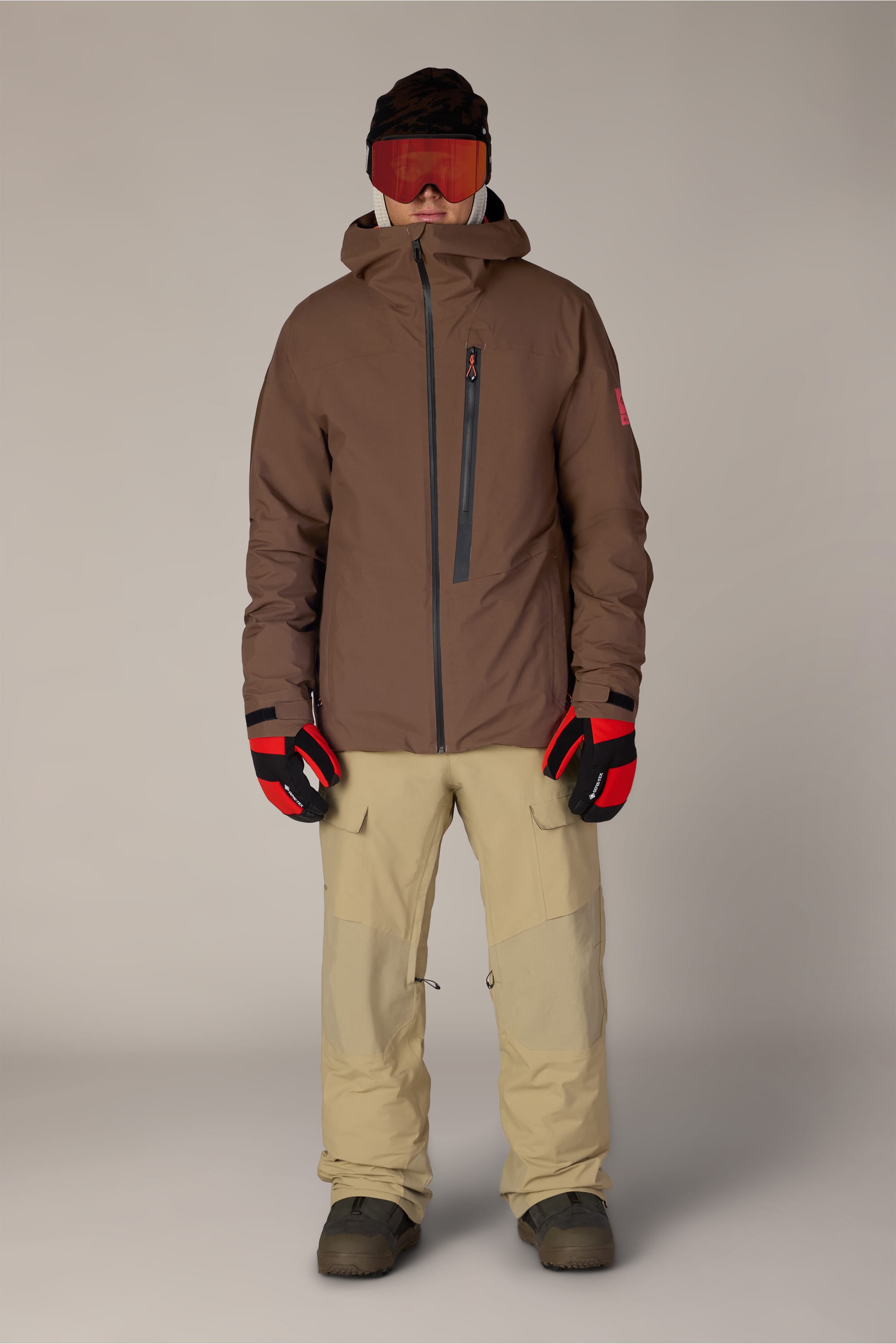 A person wearing the 686 Men’s GORE-TEX® GT Thermagraph® Jacket stands facing forward against a plain background, paired with beige snow pants, red gloves, brown boots, and red ski goggles.