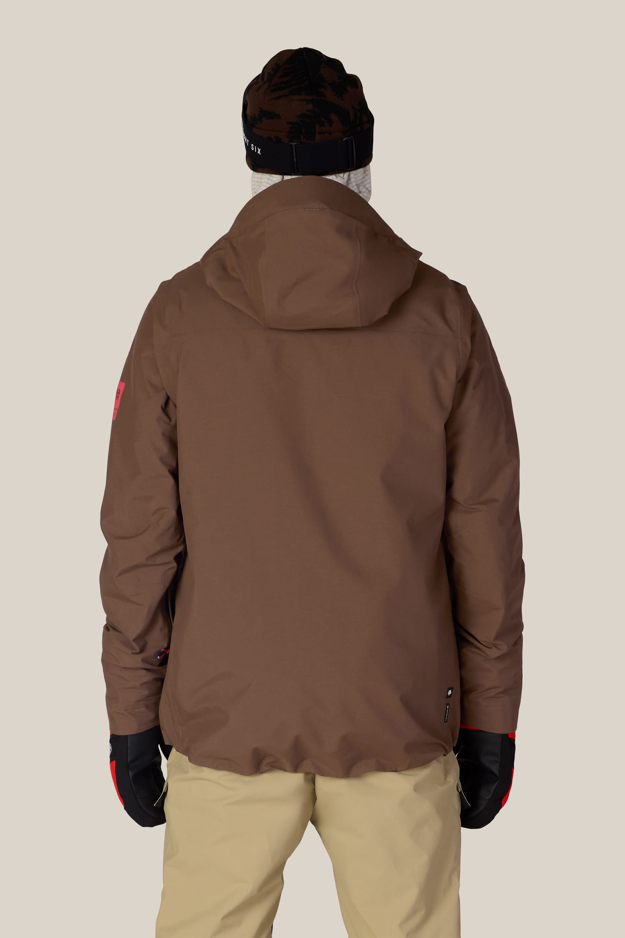 A person wearing the 686 Men’s GORE-TEX® GT Thermagraph® Jacket by 686, along with beige pants, black gloves, and a black beanie, is standing facing away from the camera against a plain background.