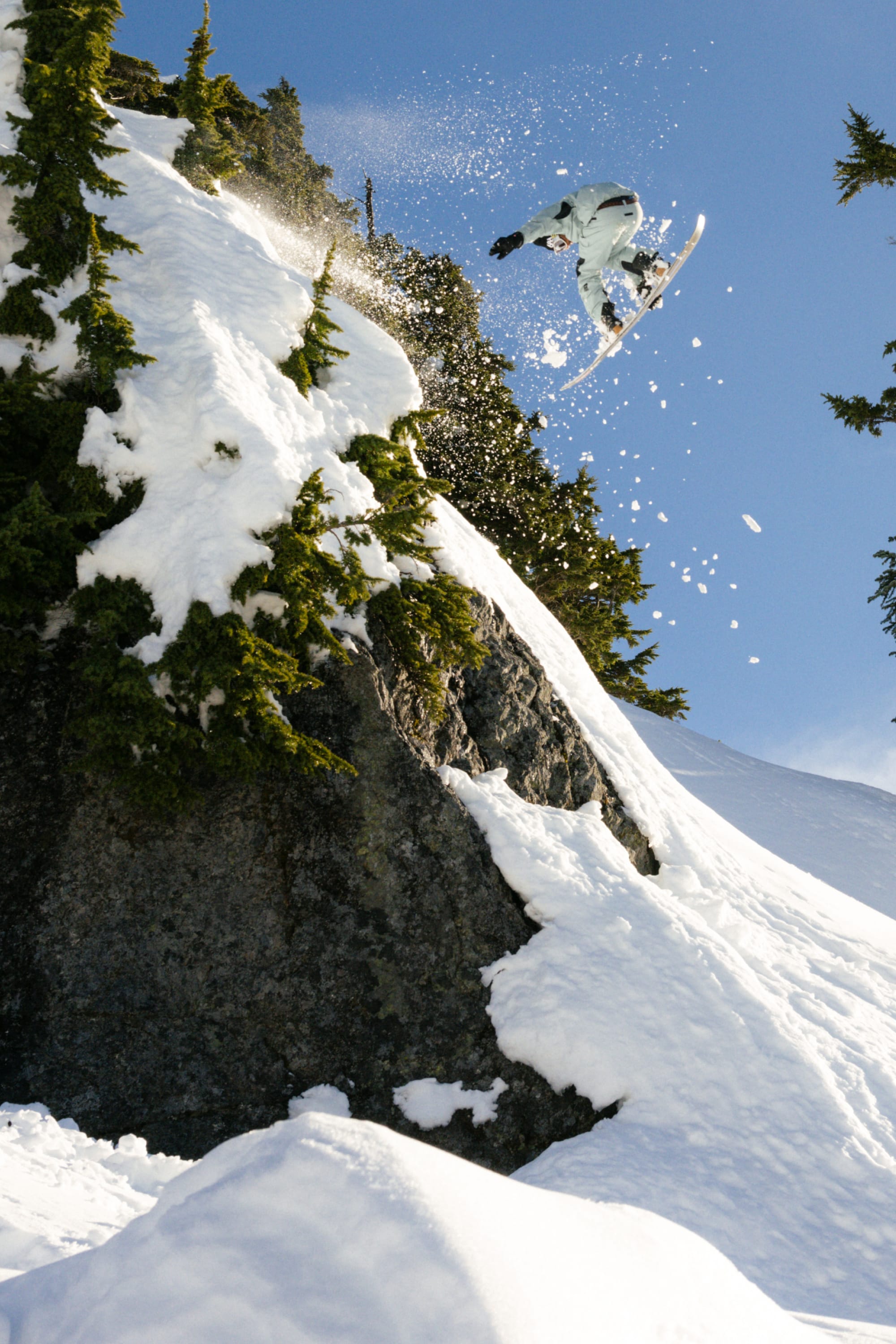 A snowboarder in the 686 Men's GORE-TEX GT Thermagraph® Jacket launches off a snowy cliff, surrounded by snow-laden trees and a clear blue sky, spraying powder behind as they soar through the air.