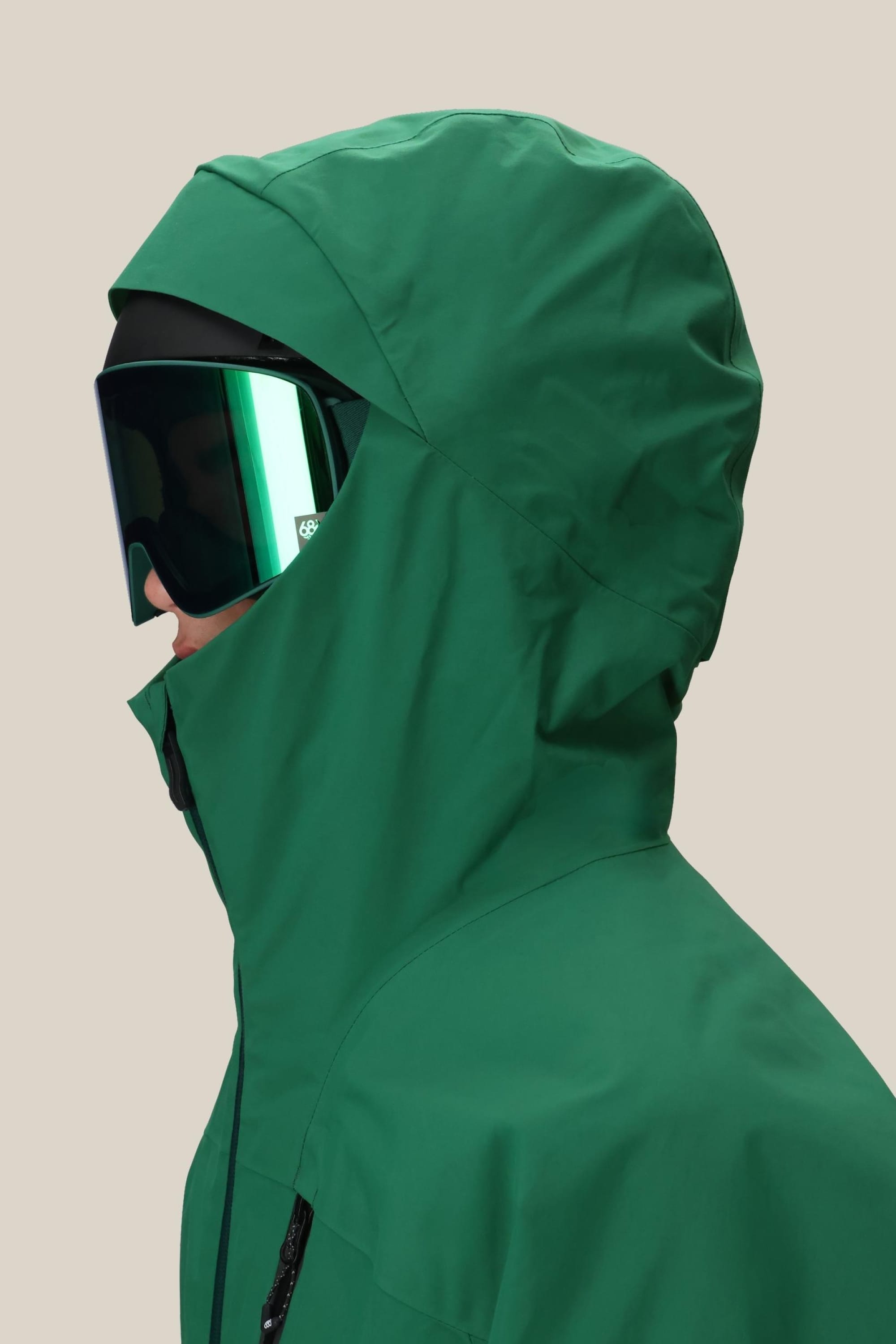 A person in a green 686 Men's GORE-TEX GT Thermagraph® Jacket with a hood and large reflective ski goggles, shown in profile against a plain beige background.