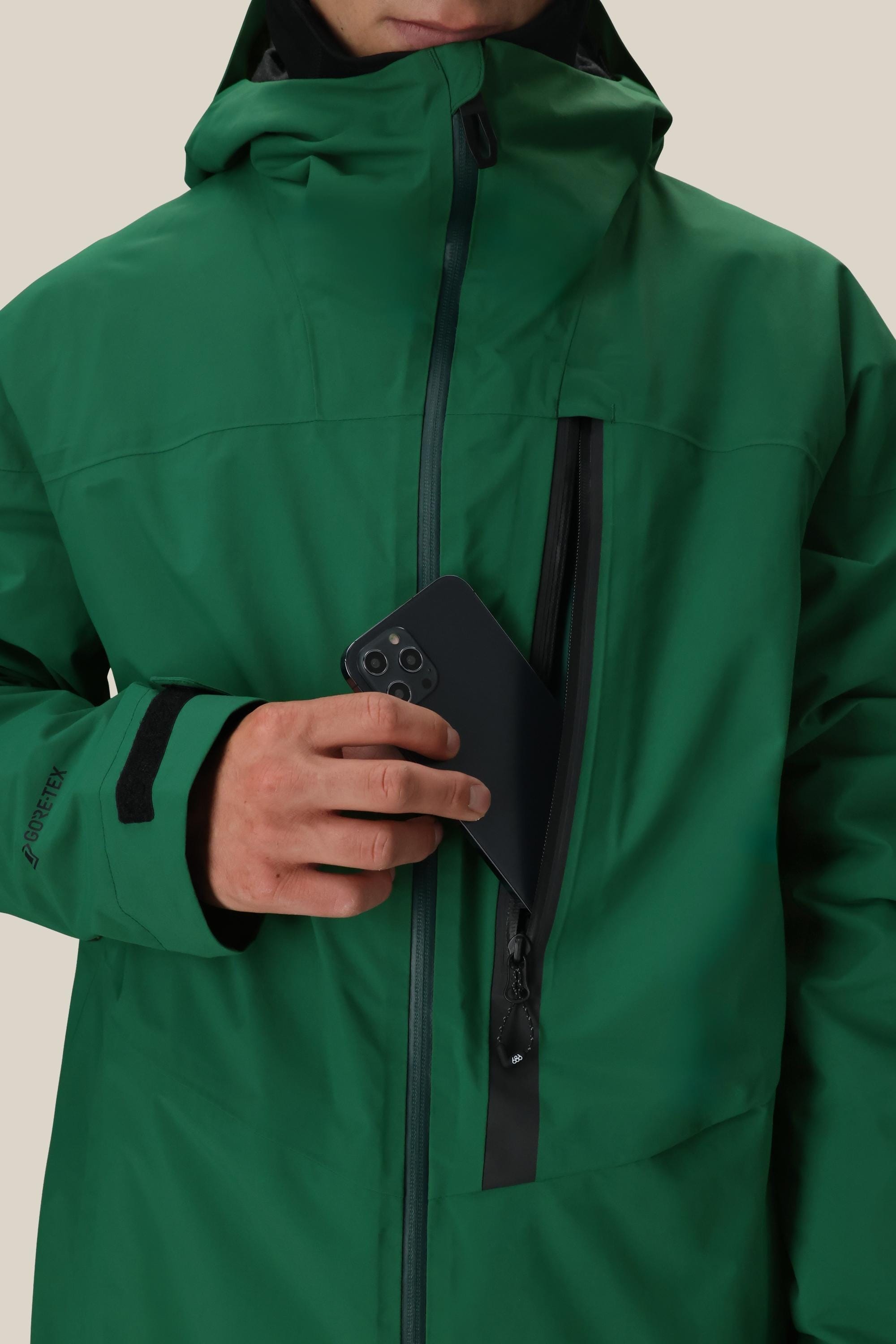 A person in a green 686 Men's GORE-TEX GT Thermagraph® Jacket with a hood partially unzips the waterproof chest pocket and puts a black smartphone inside. The 686 jacket features Velcro cuffs and is set against a plain, light background.