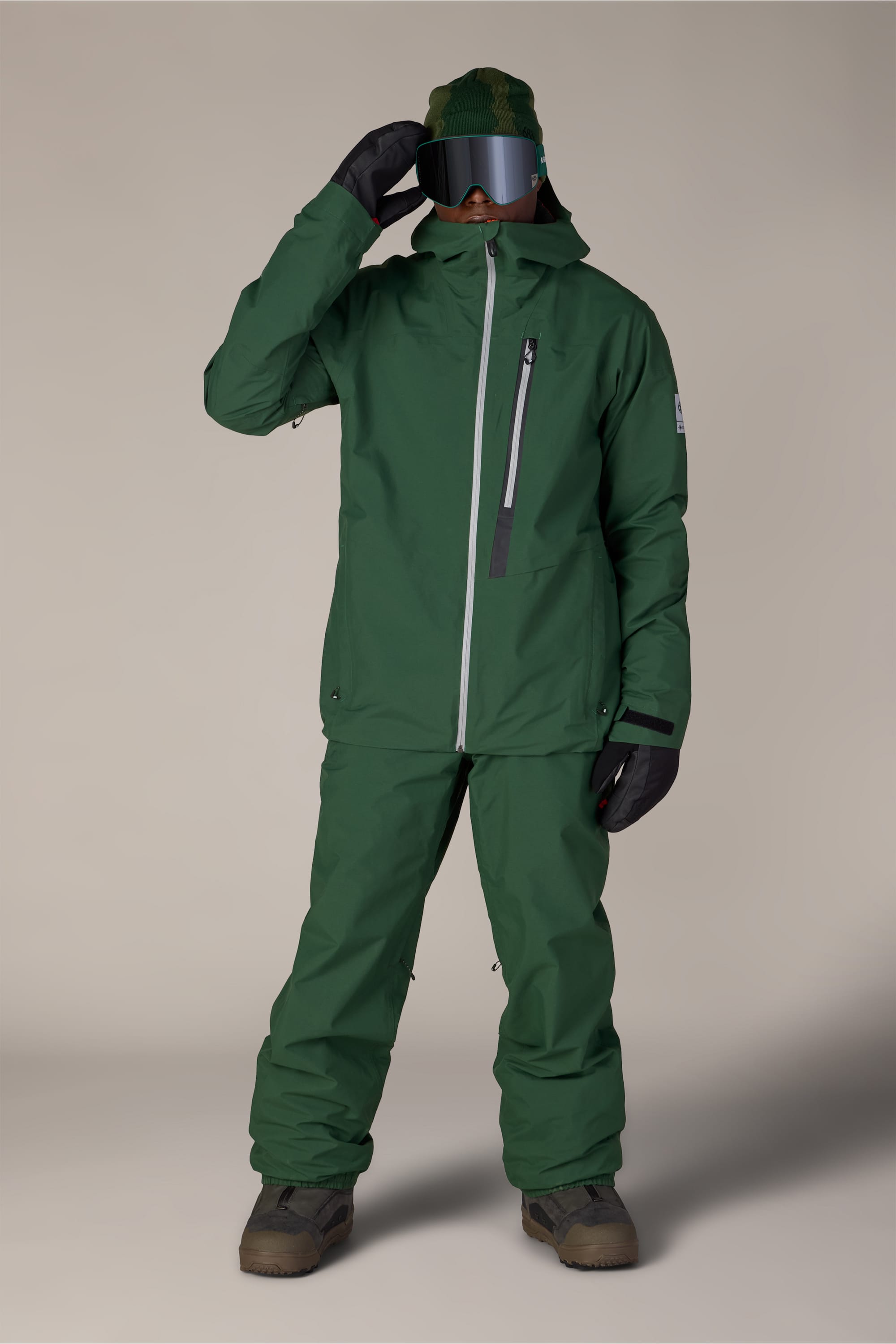 A person wearing the 686 Men’s GORE-TEX® GT Thermagraph® Jacket, matching pants, black gloves, a green beanie, and ski goggles stands on a neutral background, facing forward with one hand adjusting their goggles.