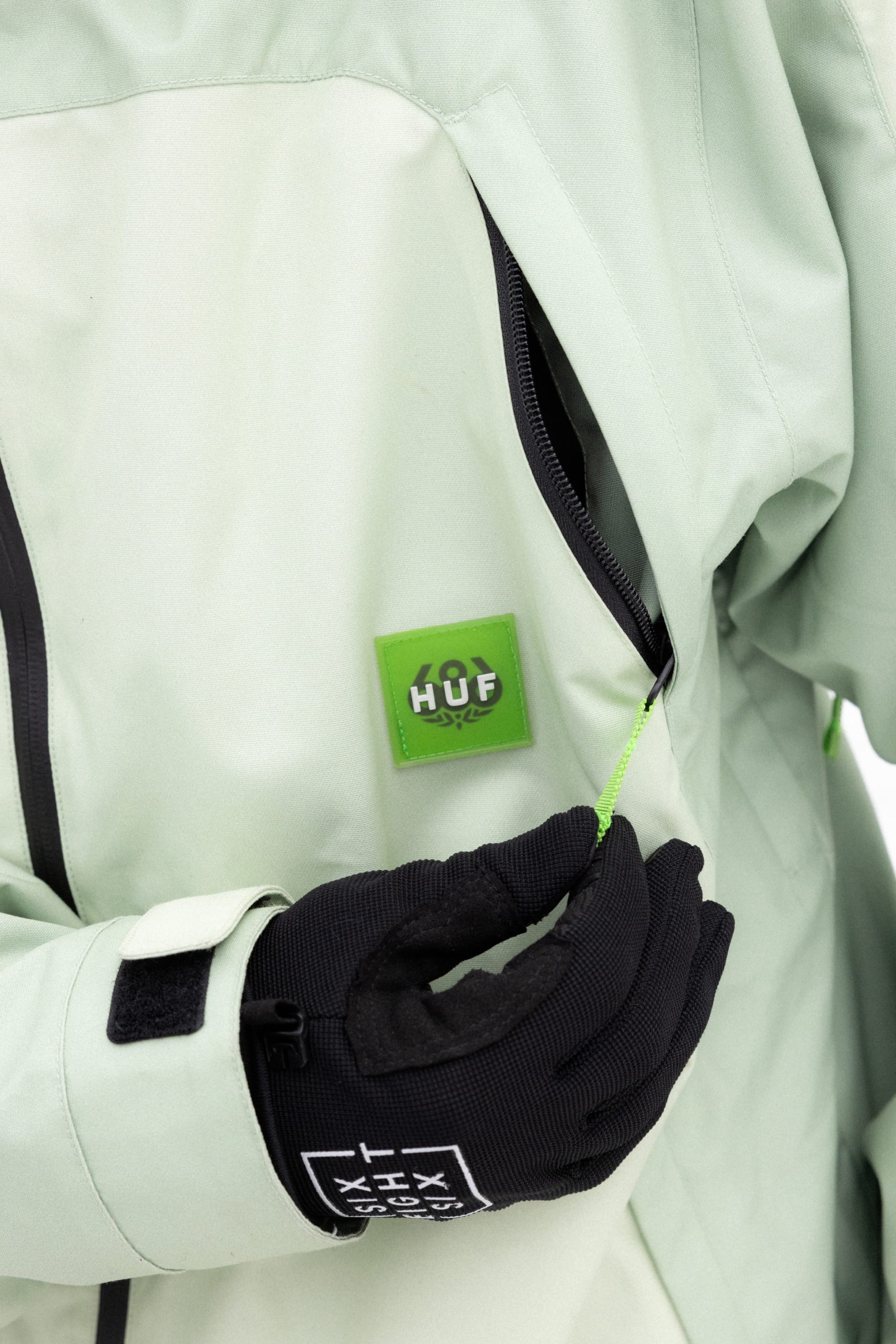 A person in a light green 686 Mens HUF Double H Shell Jacket pulls on a zipper, wearing black gloves. The chest shows the 686 logo patch on the polyester waterproof jacket.