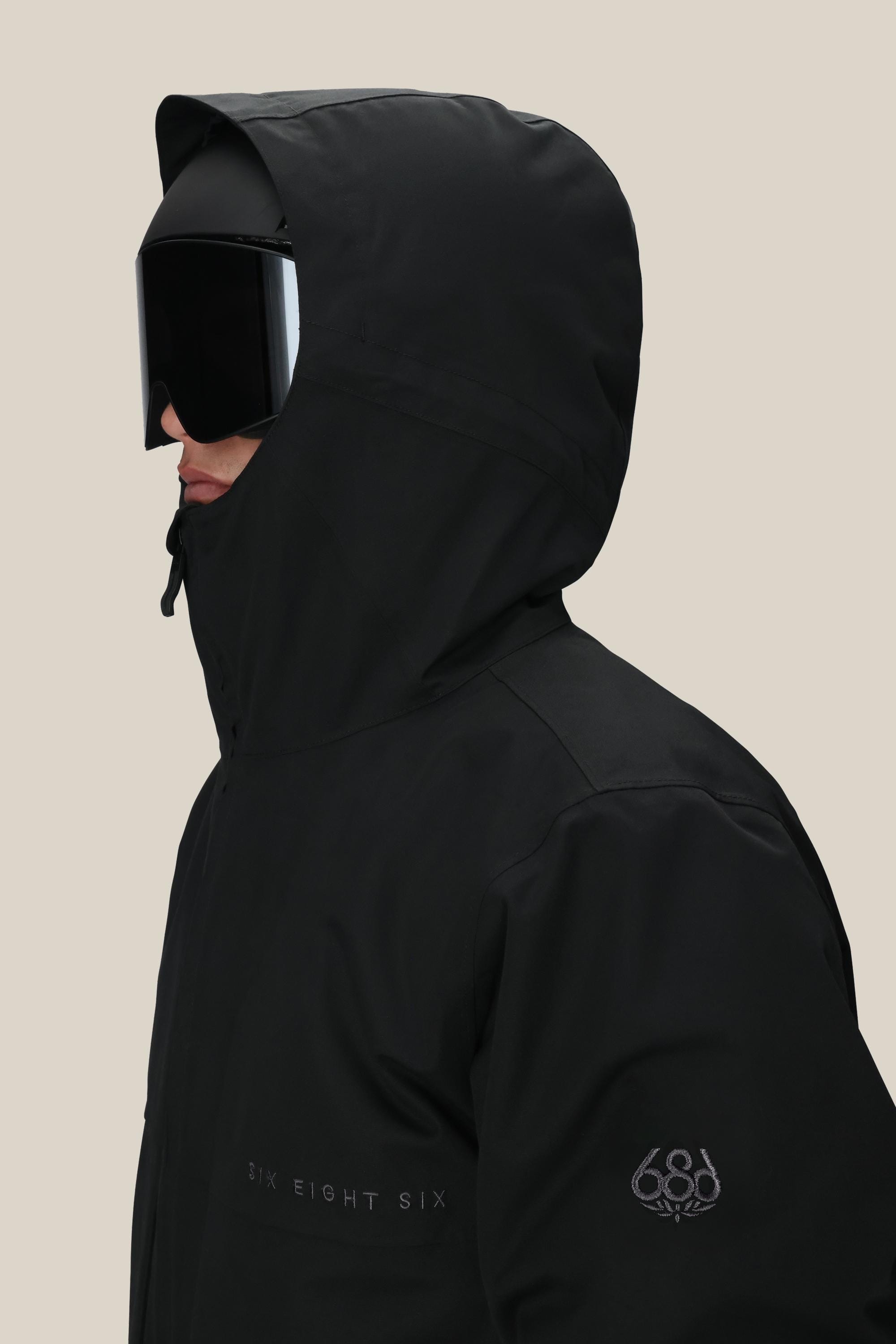 A person wearing a black 686 Men's SMARTY® 3-In-1 Form Jacket with the hood up, large black ski goggles, and visible 686 branding on the chest and sleeve.