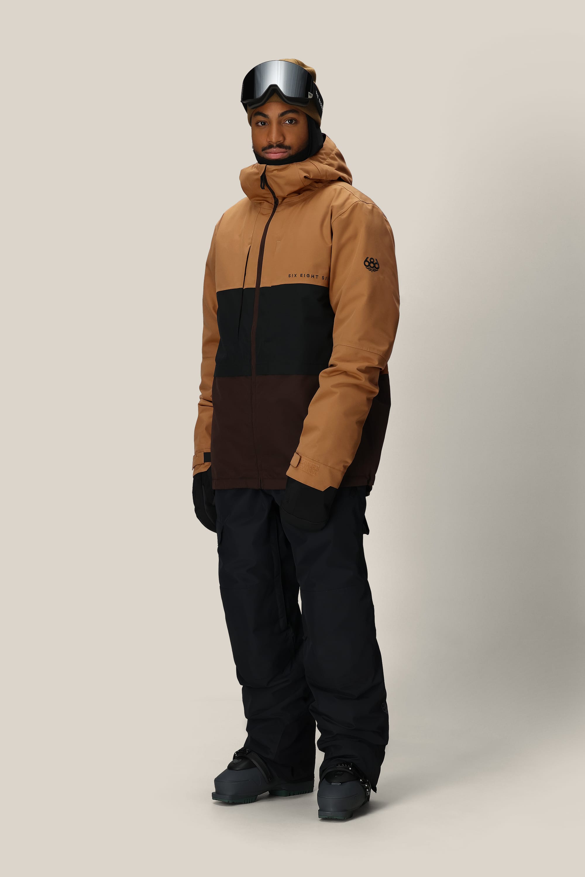 A person stands against a plain light background wearing the 686 Men's SMARTY® 3-In-1 Form Jacket in brown and black, paired with black snow pants, gloves, boots, and ski goggles resting on their forehead.